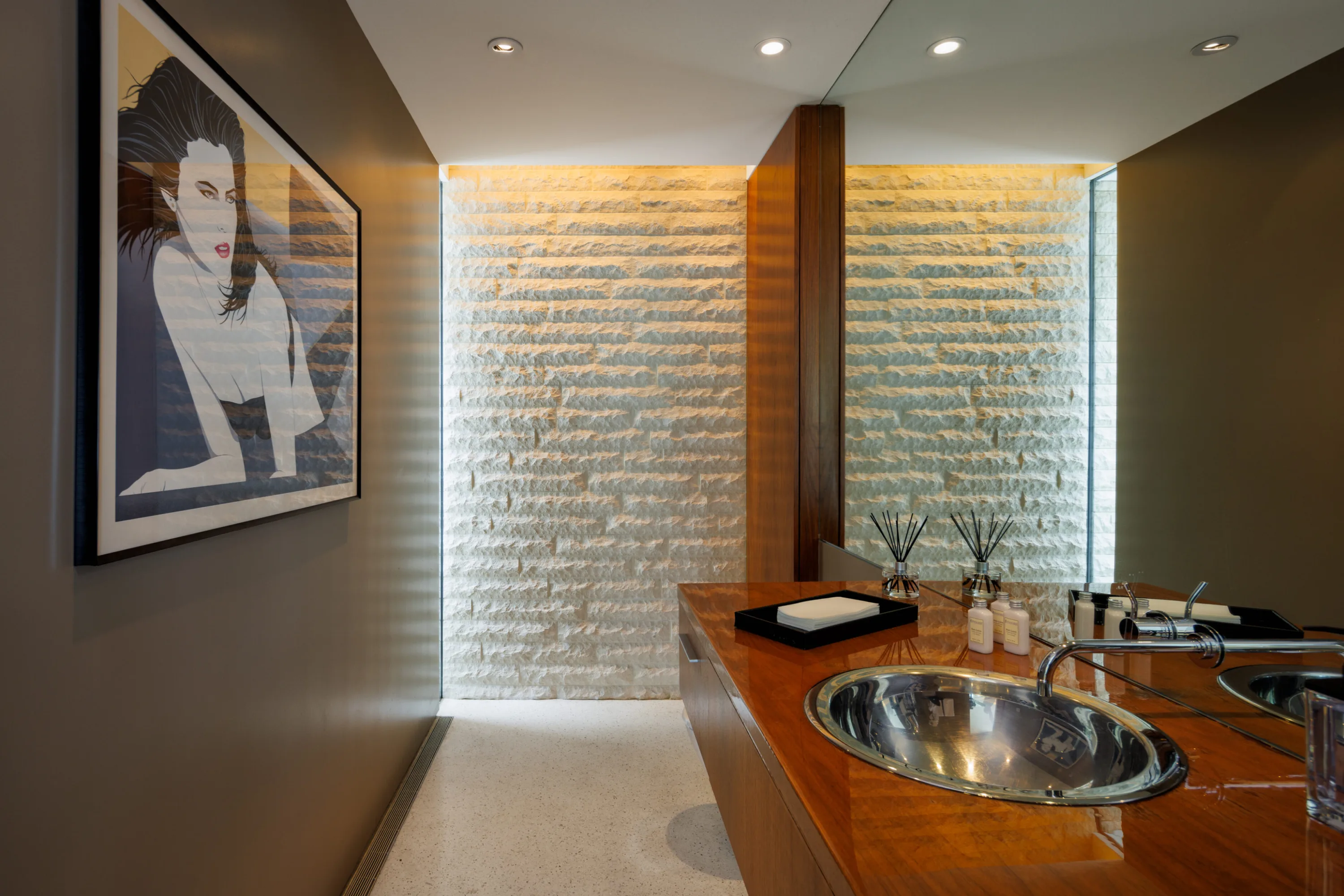Powder room with twin basins in a lacquered wood vanity, backlit stone wall and framed pop-art print