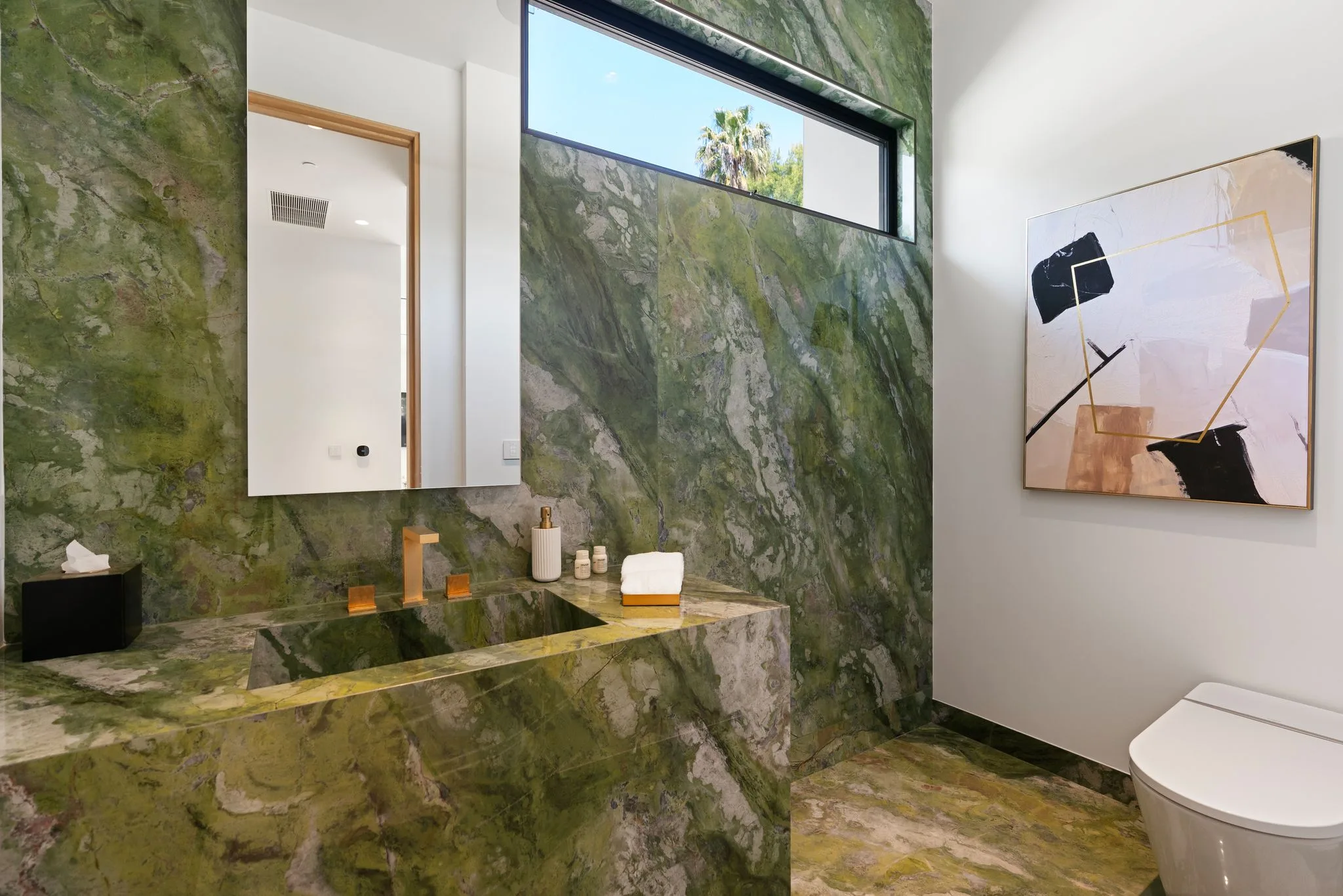 Powder room clad in dramatic green marble with a brass tap and slot window to a palm