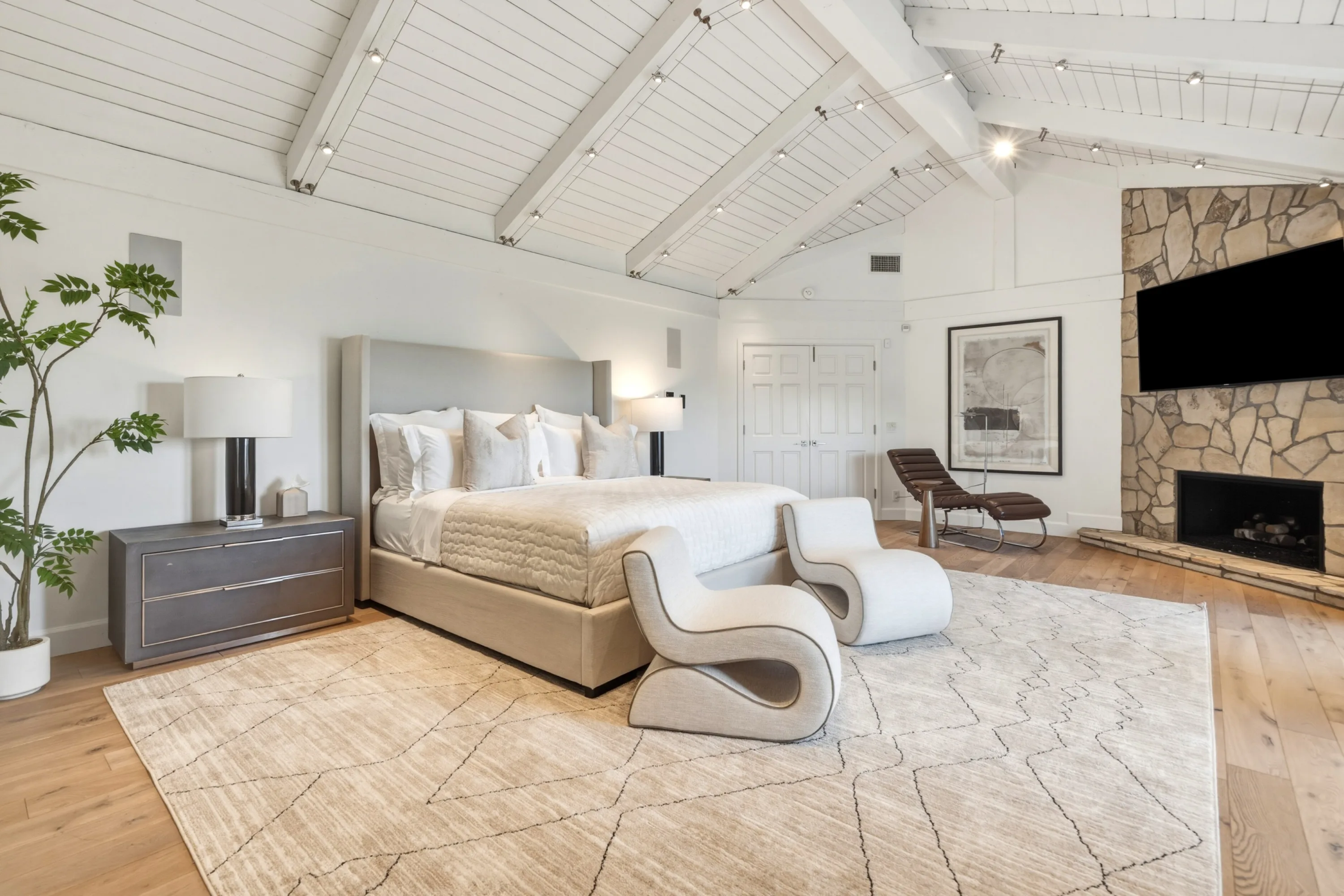Primary suite with king bed, beamed cathedral ceiling and a stone fireplace beneath the wall TV