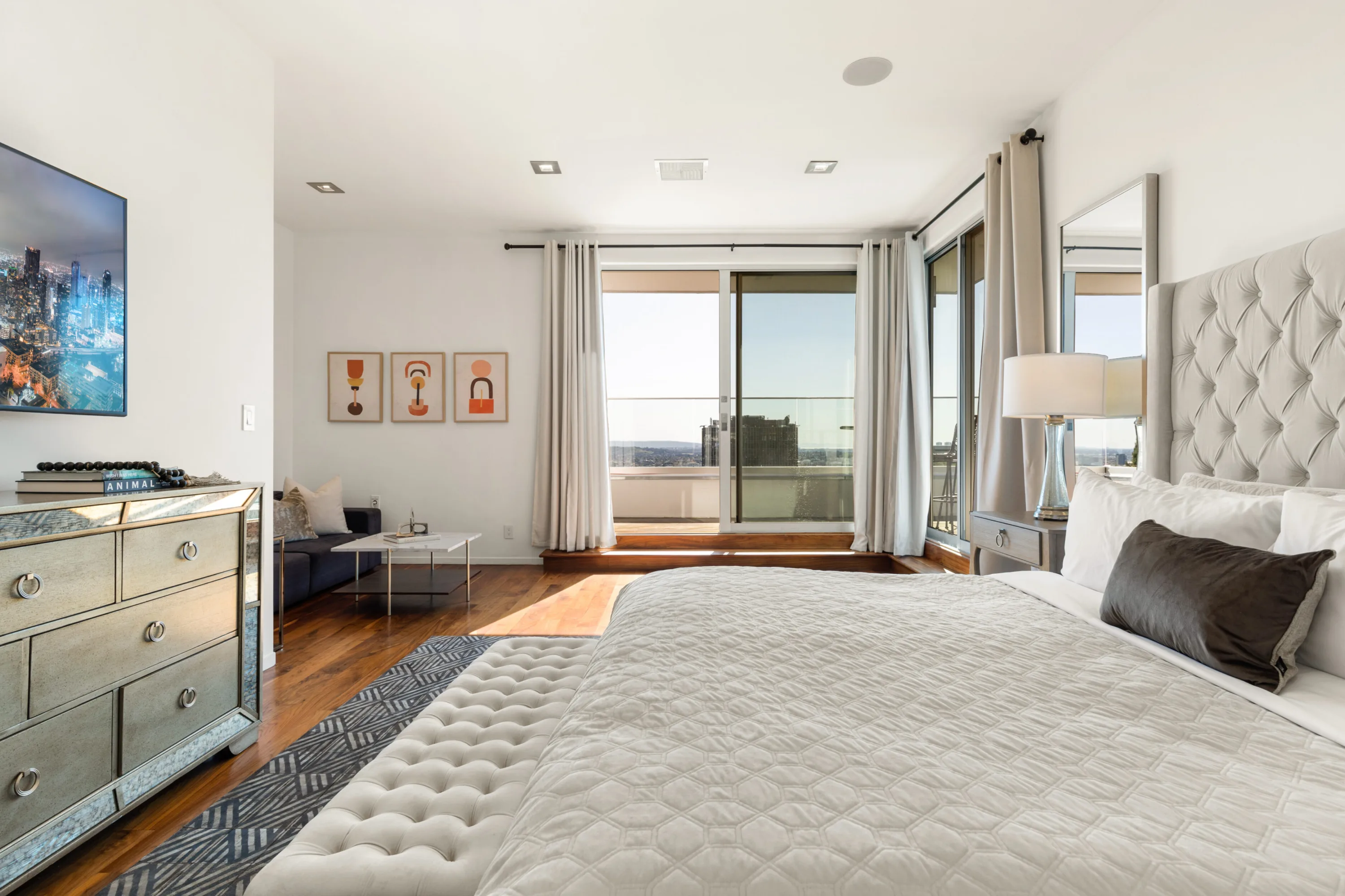 Spacious bedroom with mirrored dresser, sofa nook and balcony framing the downtown high-rise view