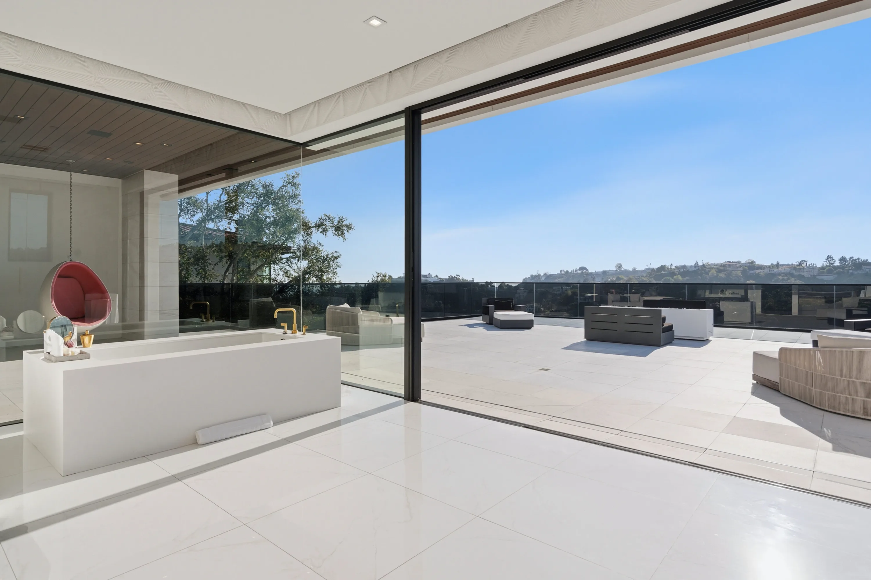 Primary bath's freestanding soaking tub opening through retracted glass walls onto a sunlit terrace above the hills