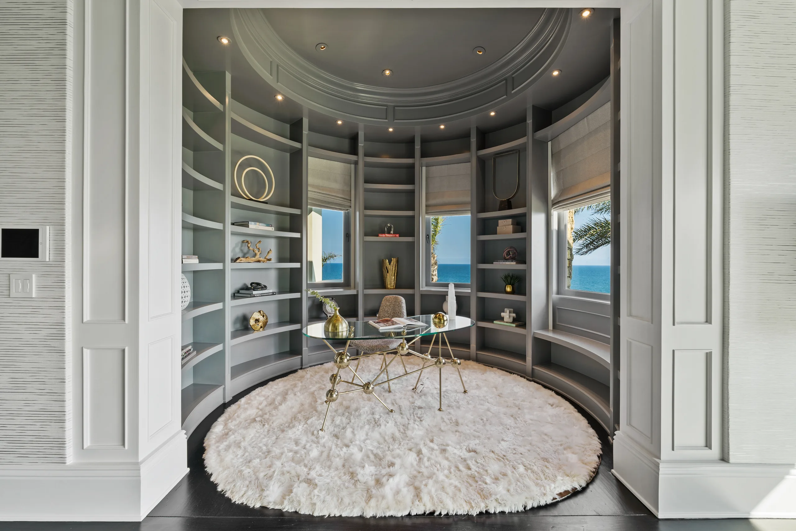 Domed study with curved grey shelving, glass desk on a shaggy round rug and blue horizon in the windows