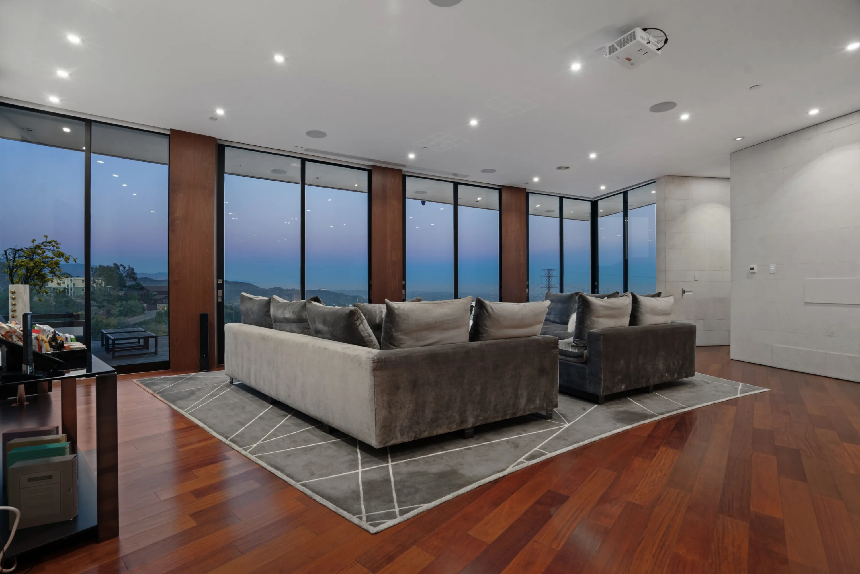 Screening lounge at dusk, plush sectional on a grey rug facing the ceiling projector and twilight glass wall