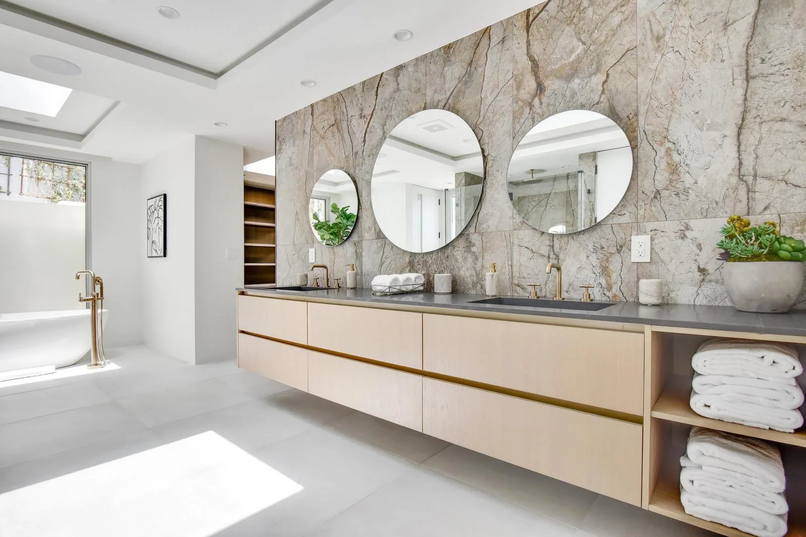 Floating oak double vanity set against a book-matched stone wall with three round mirrors and stacked towels