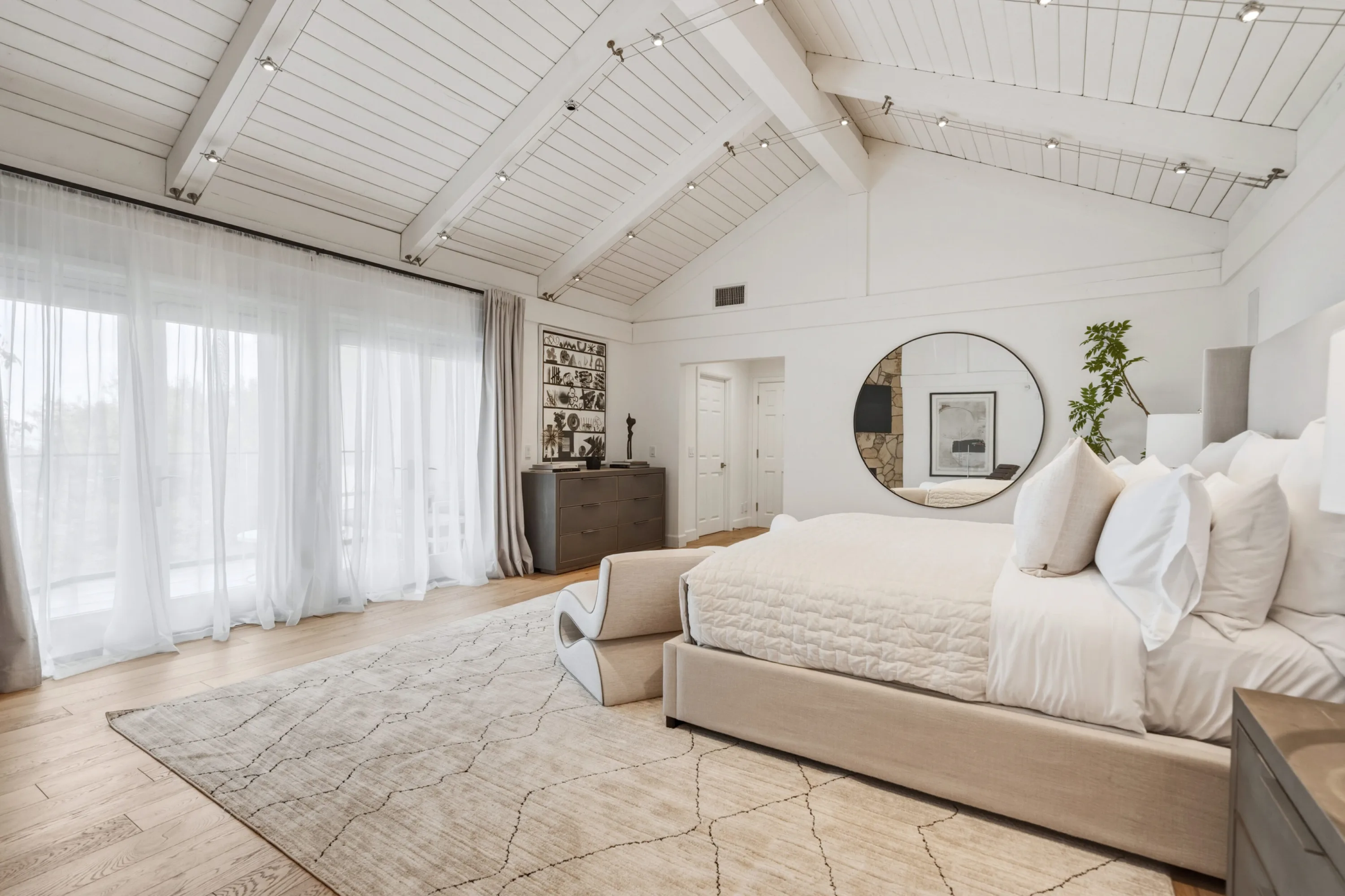 Sheer-curtained wall of glass beside the upholstered bed in the airy vaulted main bedroom