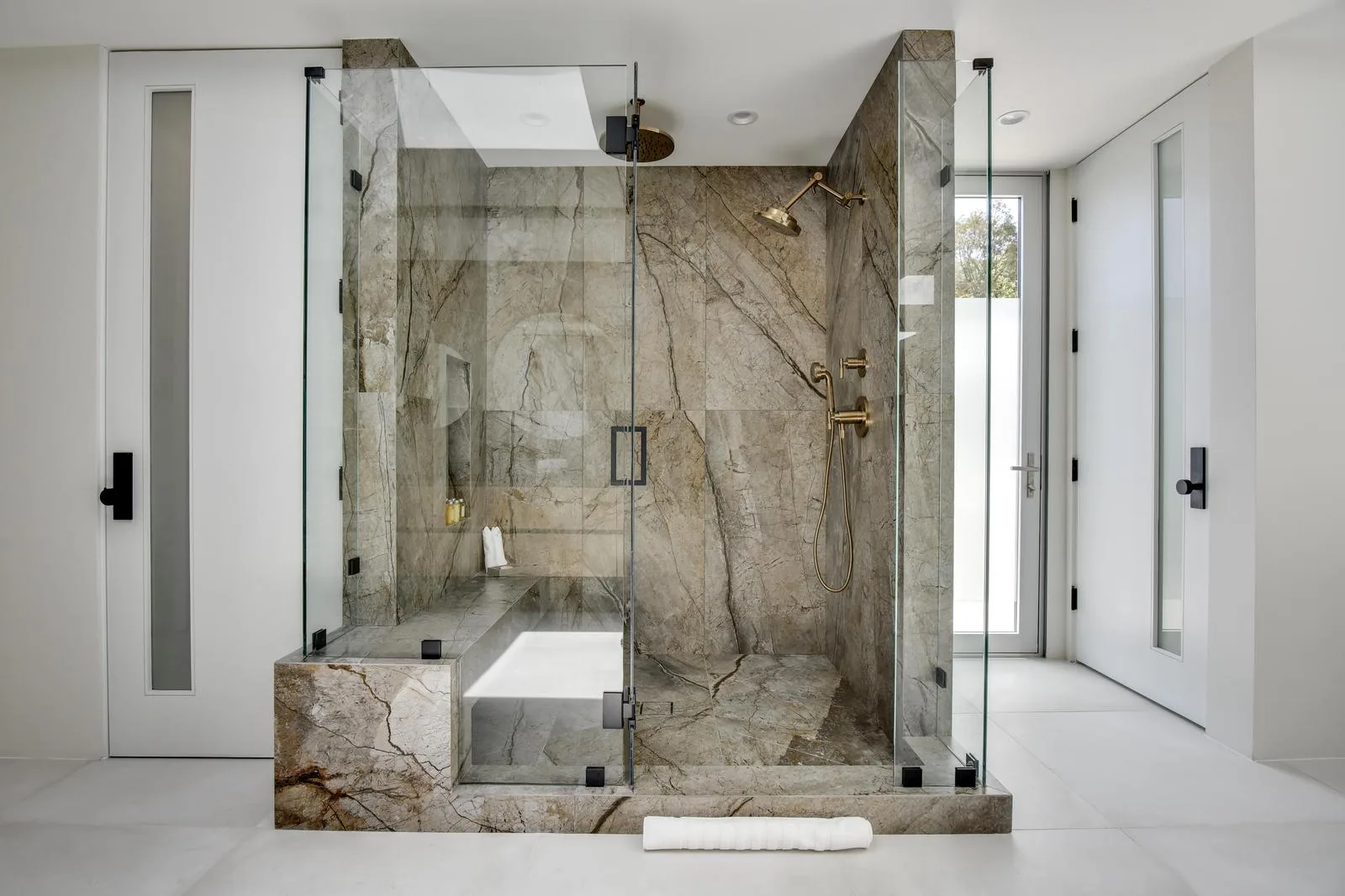 Glass-walled steam shower clad in veined stone, with brass rainhead, bench seat and overhead skylight