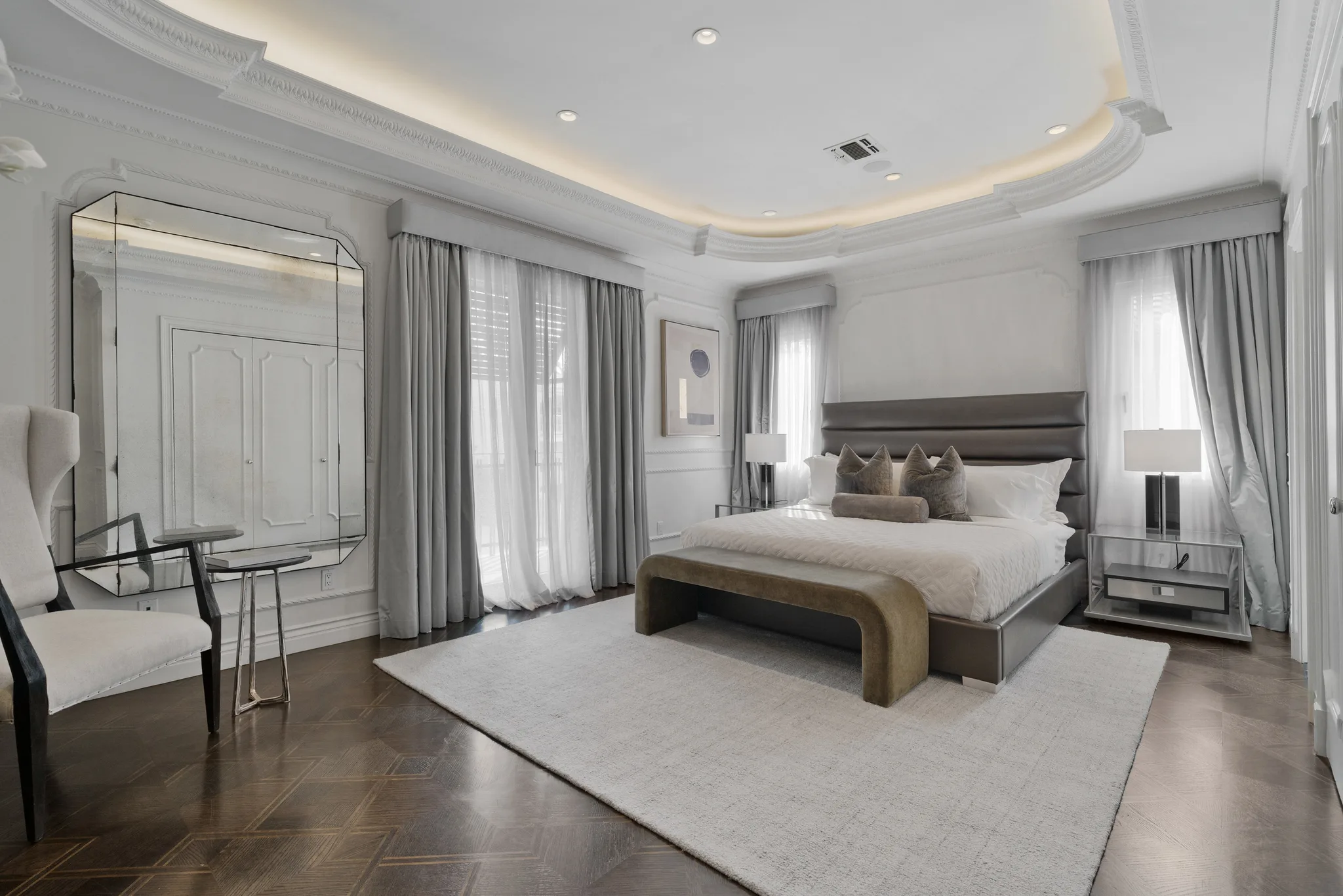 Primary suite under a coved, backlit ceiling, with leather-panelled bed, oversized mirror and herringbone wood floors