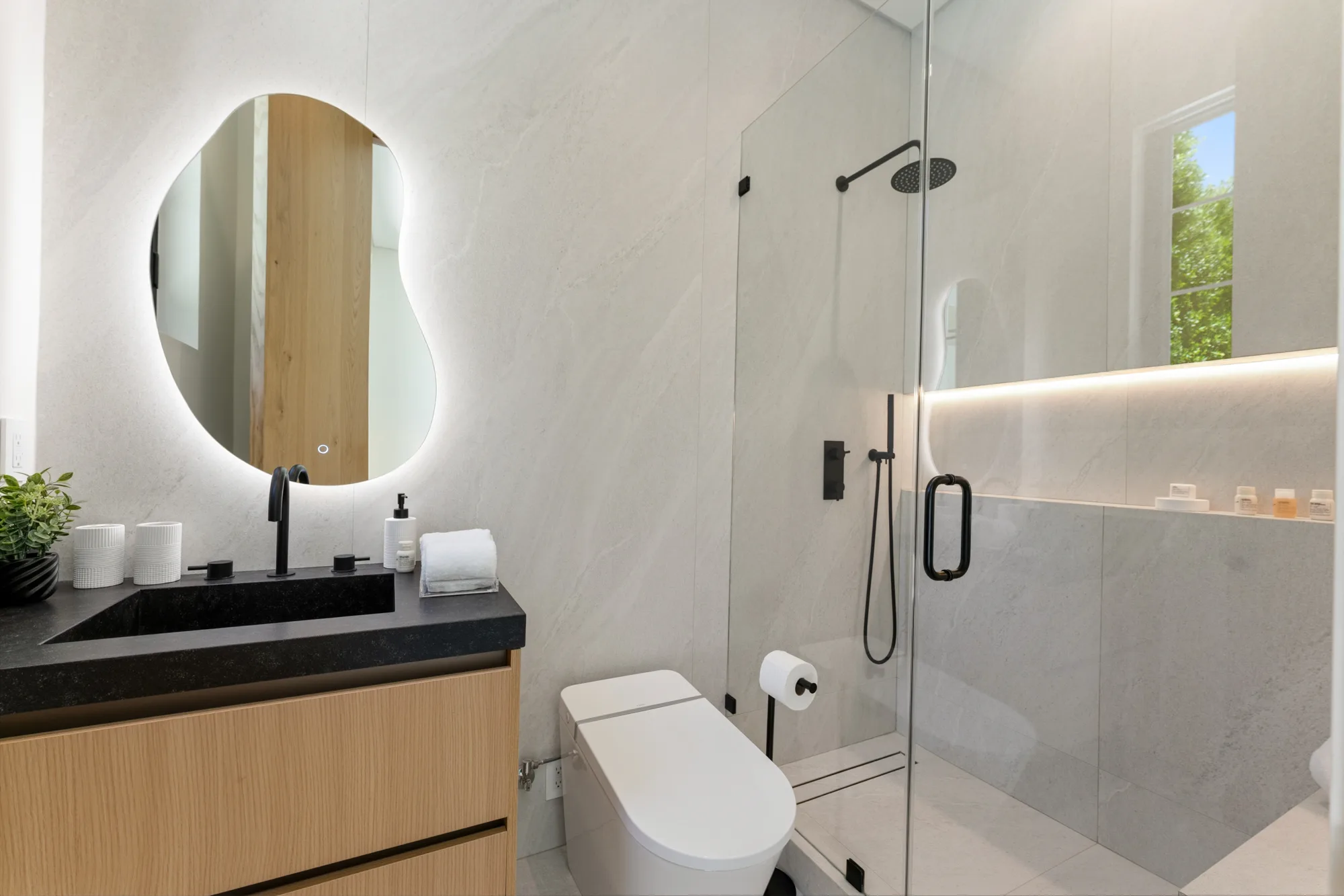 Guest bathroom with backlit pebble-shaped mirror, honed black stone sink and a glass walk-in rain shower