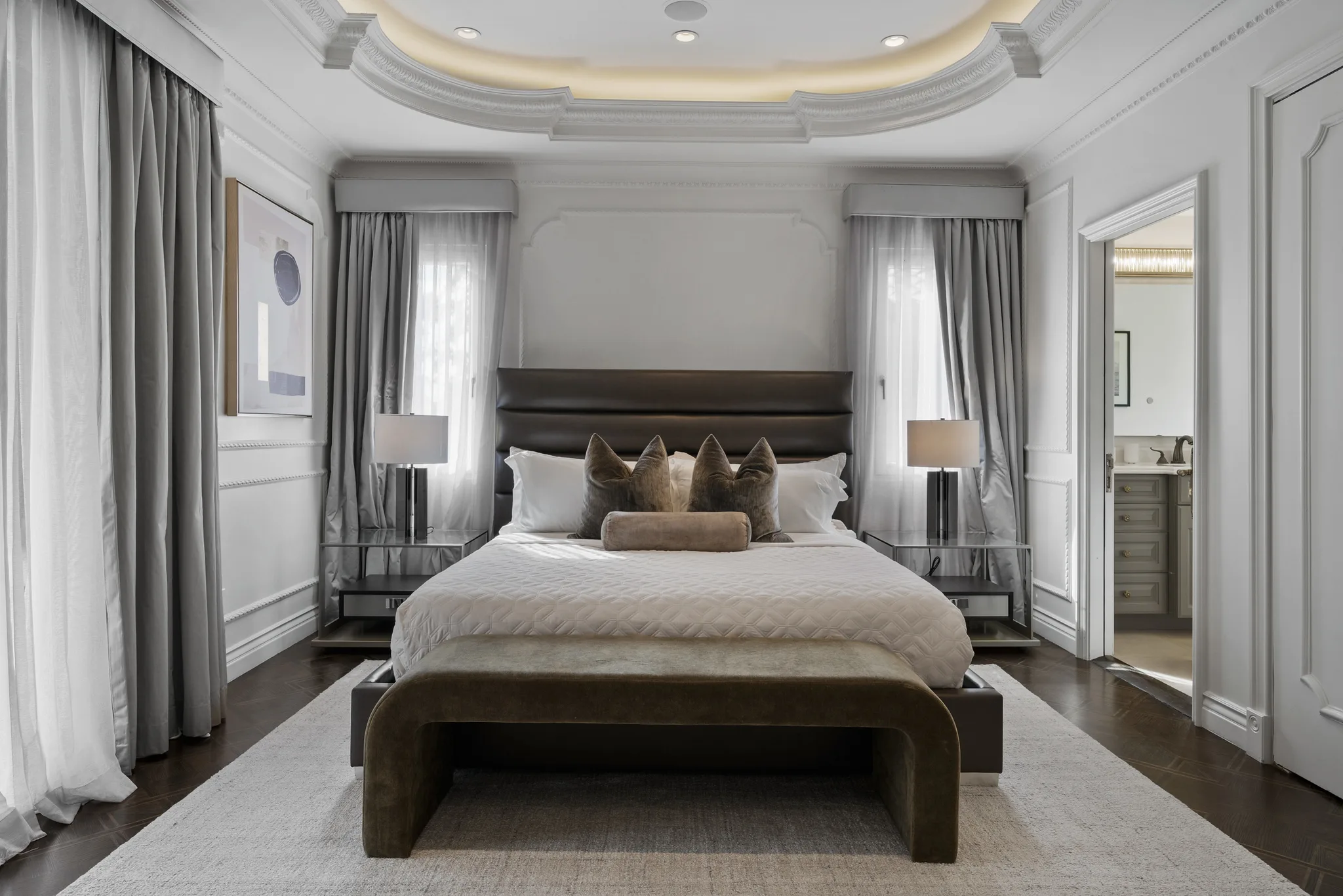 Primary bedroom head-on: tall leather headboard framed by curtained windows, velvet bench and open door to the ensuite
