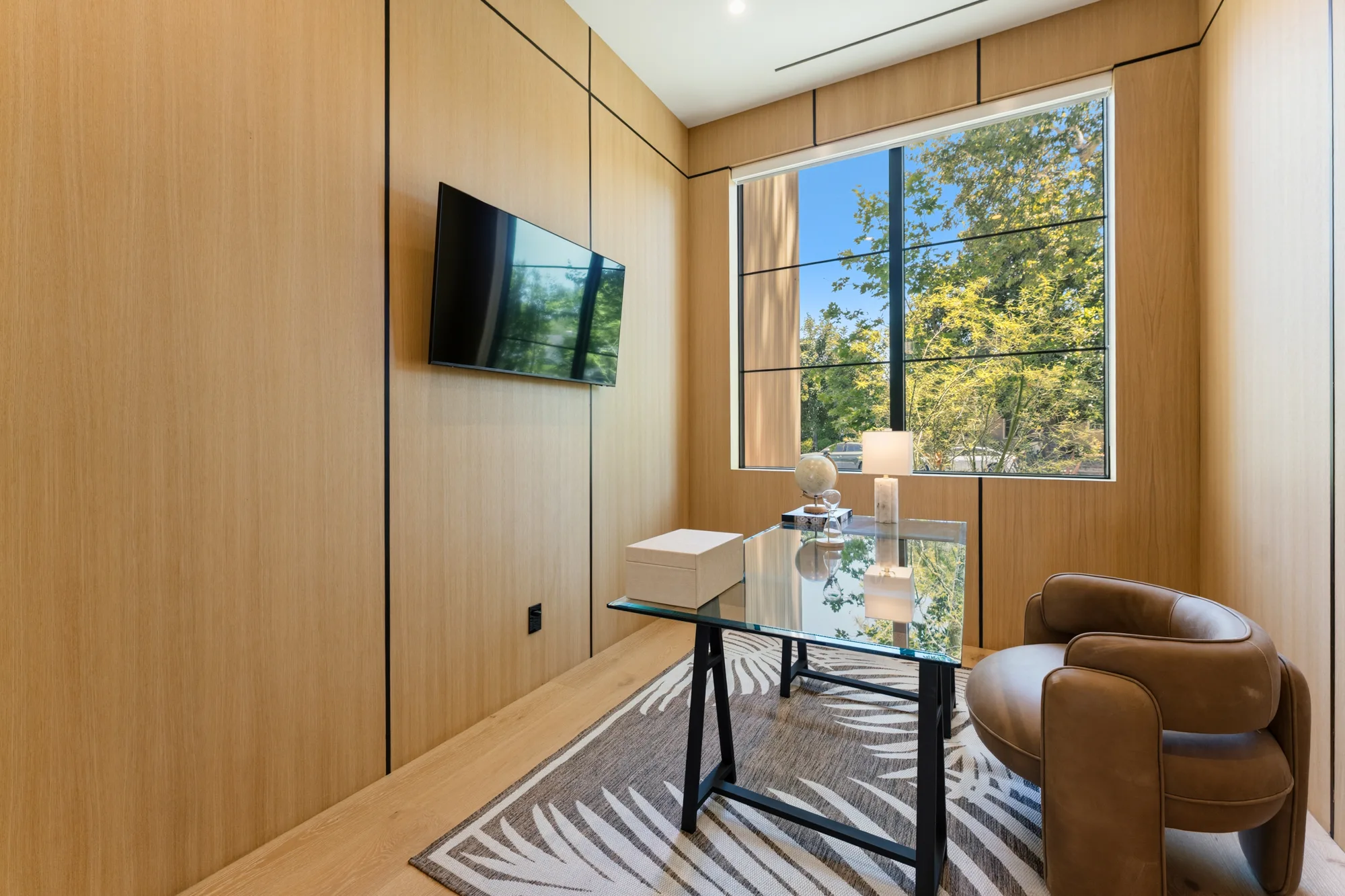 Oak-panelled study with glass desk, tan leather swivel chair and a window full of sunlit foliage