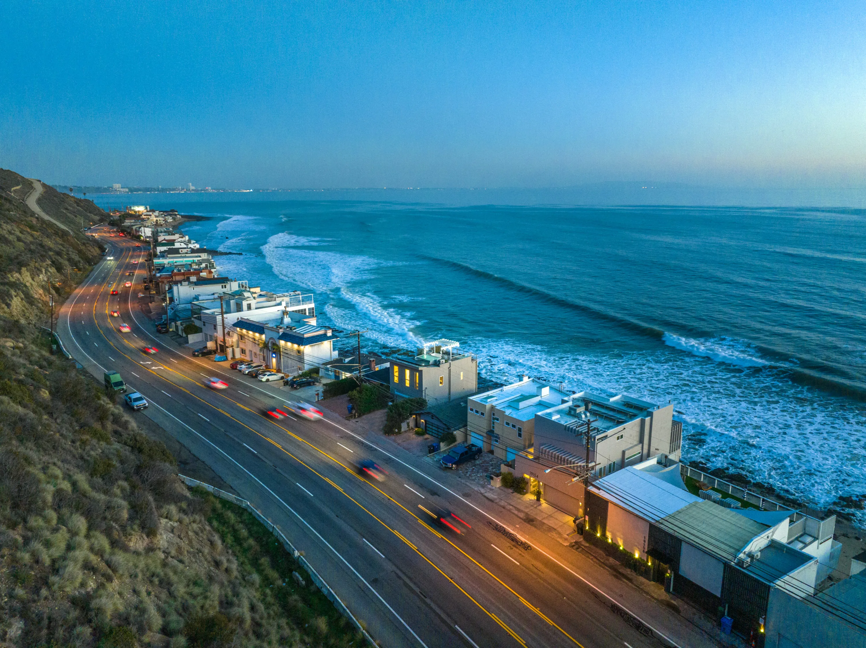 Aerial dusk view of Pacific Coast Highway curving past beach houses, headlight trails beside breaking surf