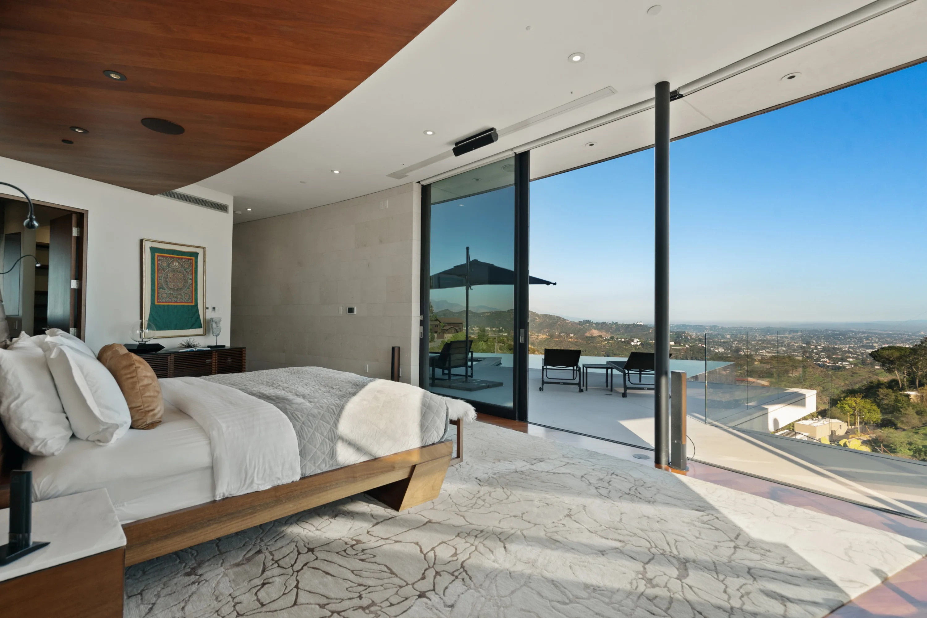Bed facing open sliders onto a terrace with parasol and loungers, hillside rooftops beyond the glass rail