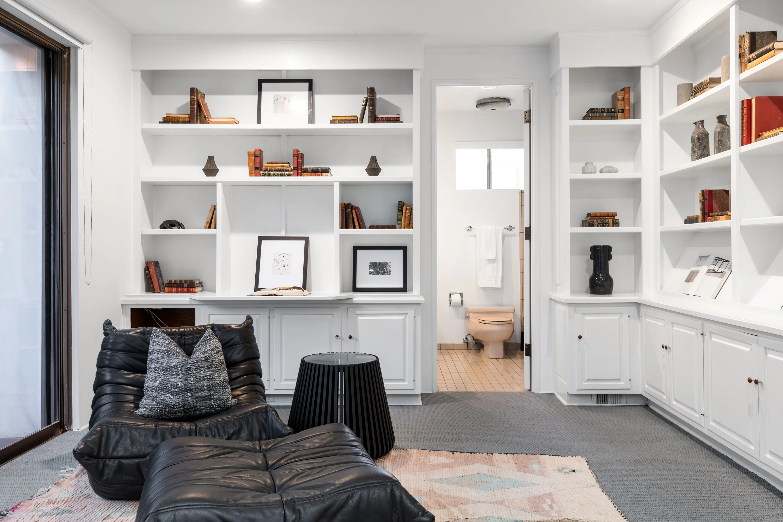 Sitting area lined with built-in white bookshelves, low leather lounger and adjoining powder room