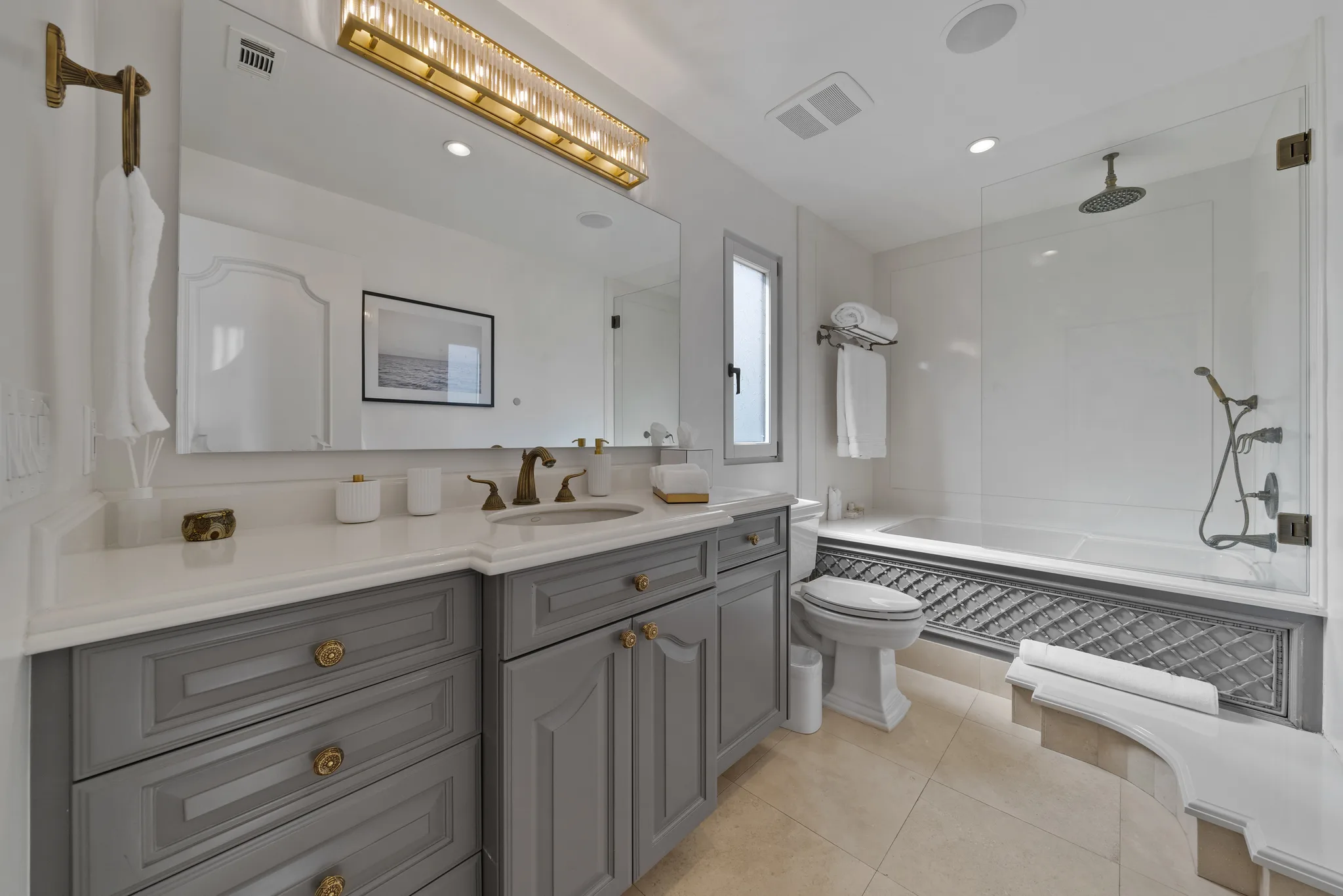 Marble-topped bathroom with brass hardware, long mirror under a fluted gold light bar and a tub beneath a rain shower