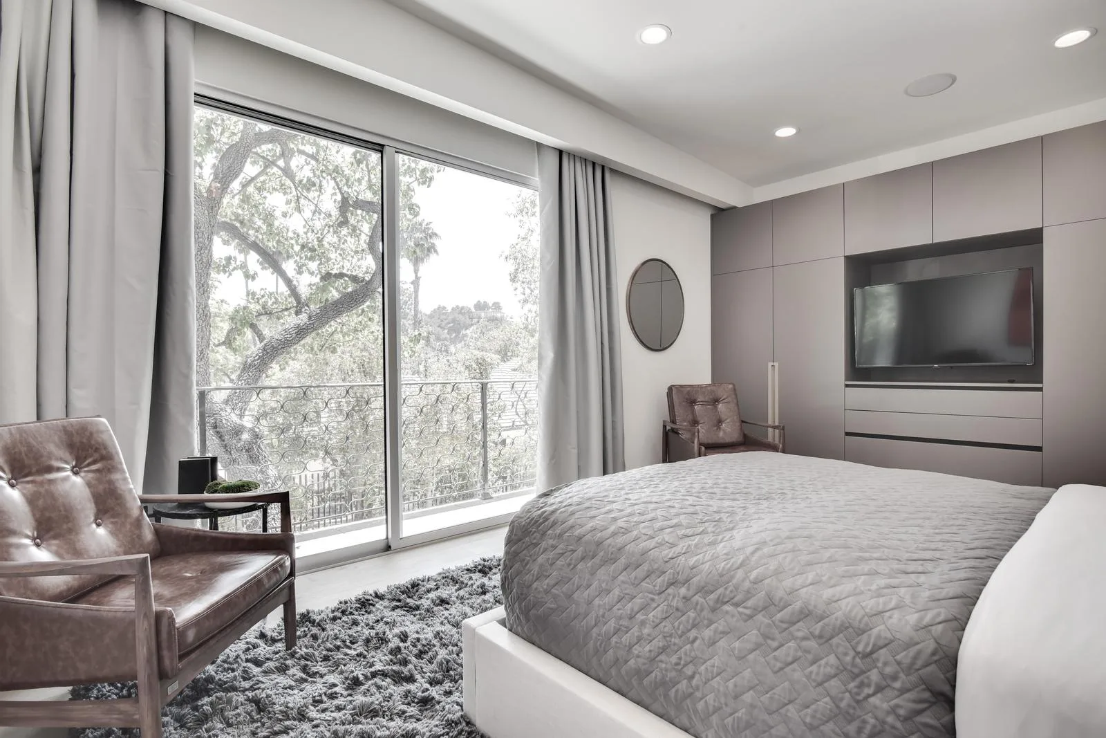 Guest bedroom in soft greys, sliding doors to a balcony framed by a mature oak, wall-mounted TV opposite the bed