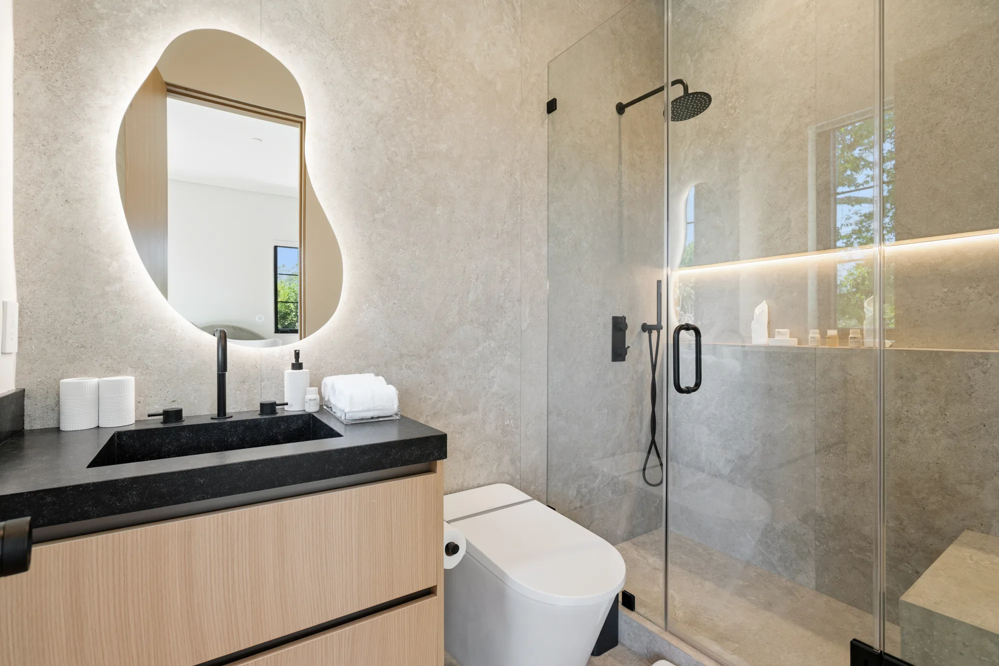 Stone-clad ensuite with illuminated organic mirror and a walk-in shower lit by a recessed niche