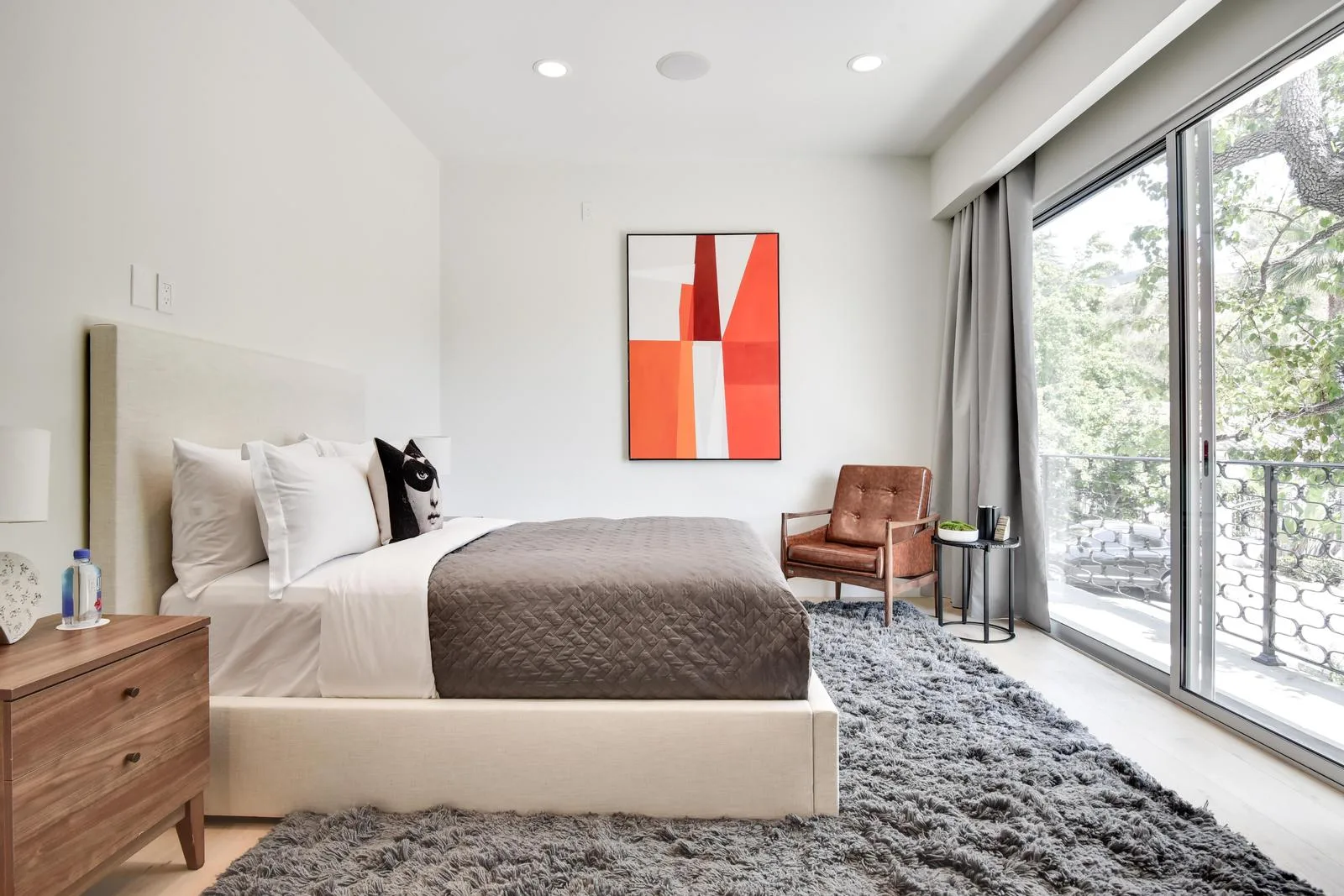 Bright bedroom with bold red-and-orange artwork, leather armchair and sliding glass doors onto a treetop balcony
