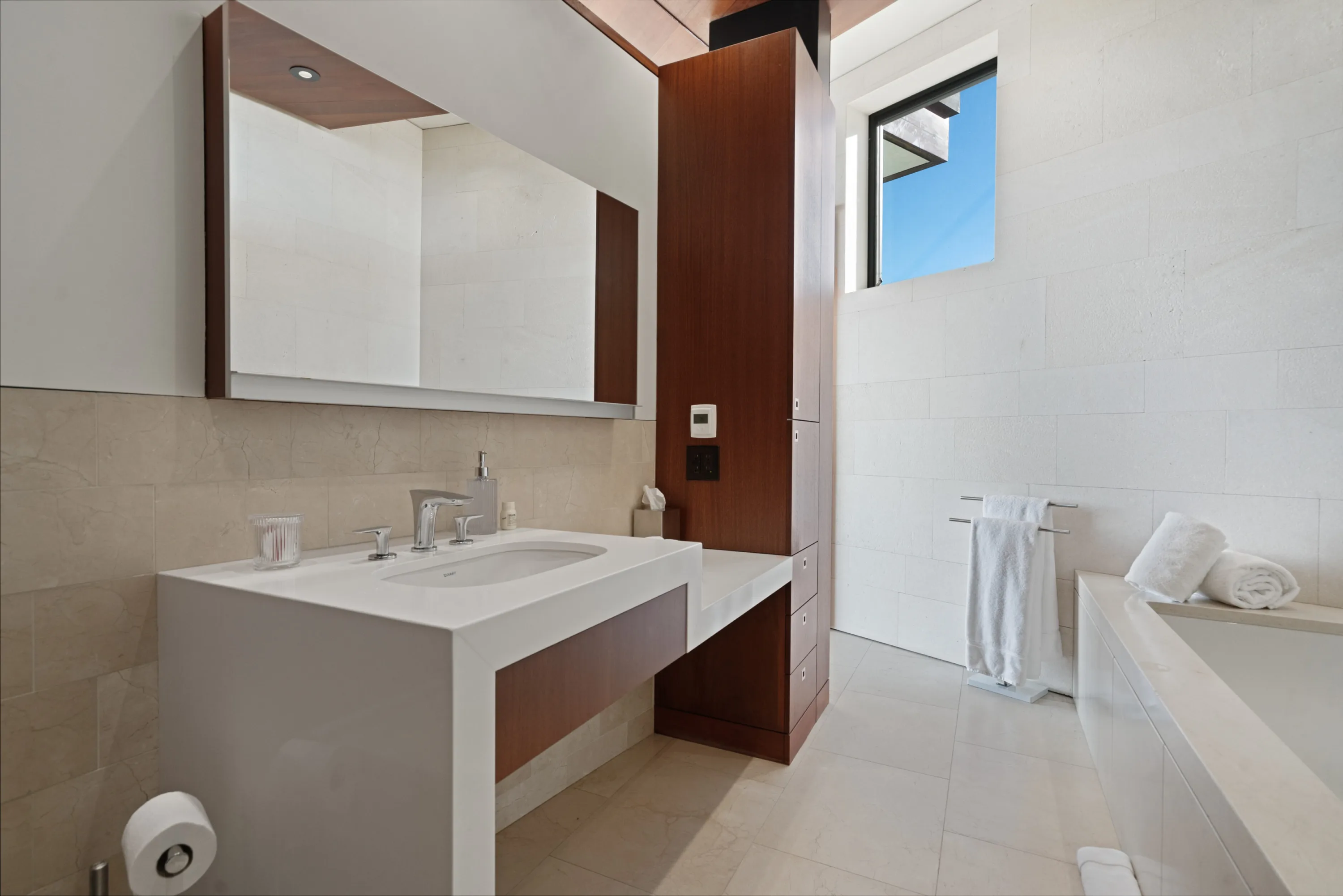 Guest bathroom in limestone and rosewood, wide mirror, tub and folded towels beneath a clerestory window
