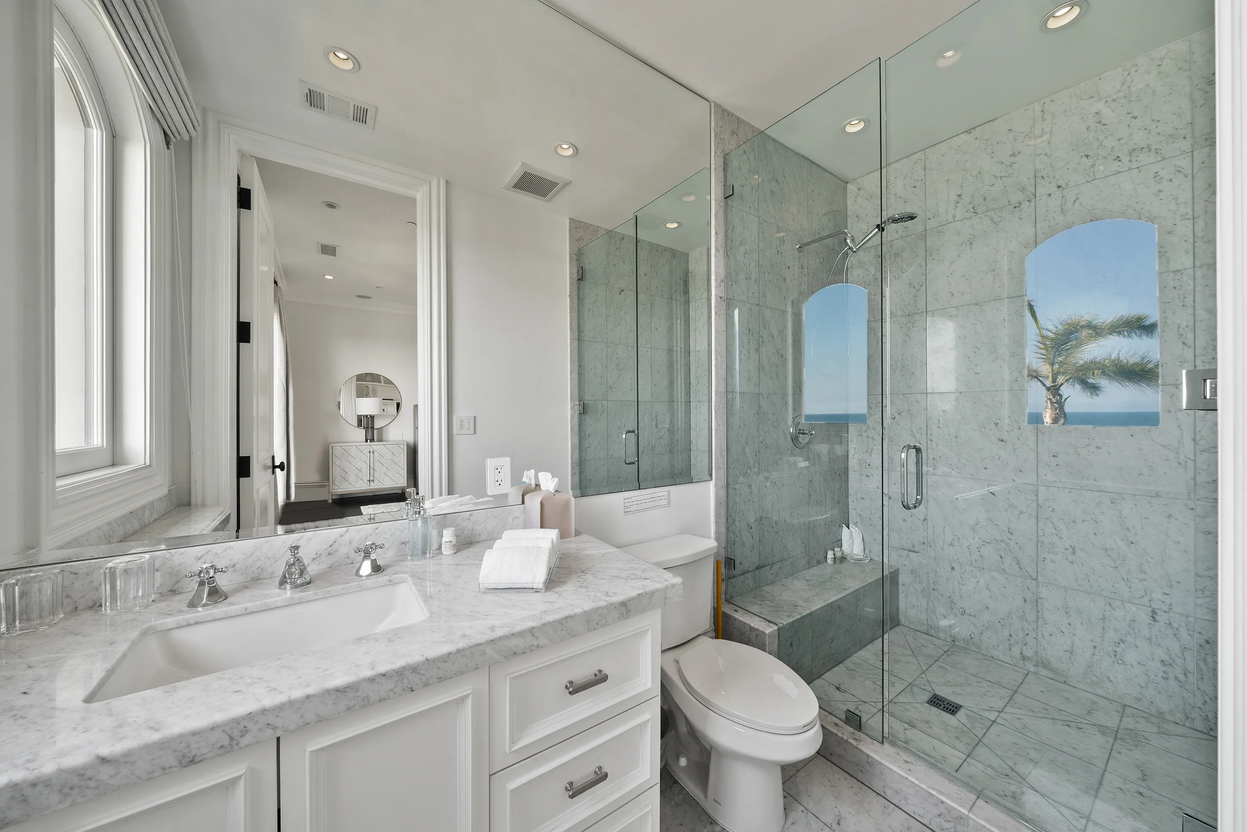 Guest bath in Carrara marble with walk-in shower and arched windows catching palm fronds and sea