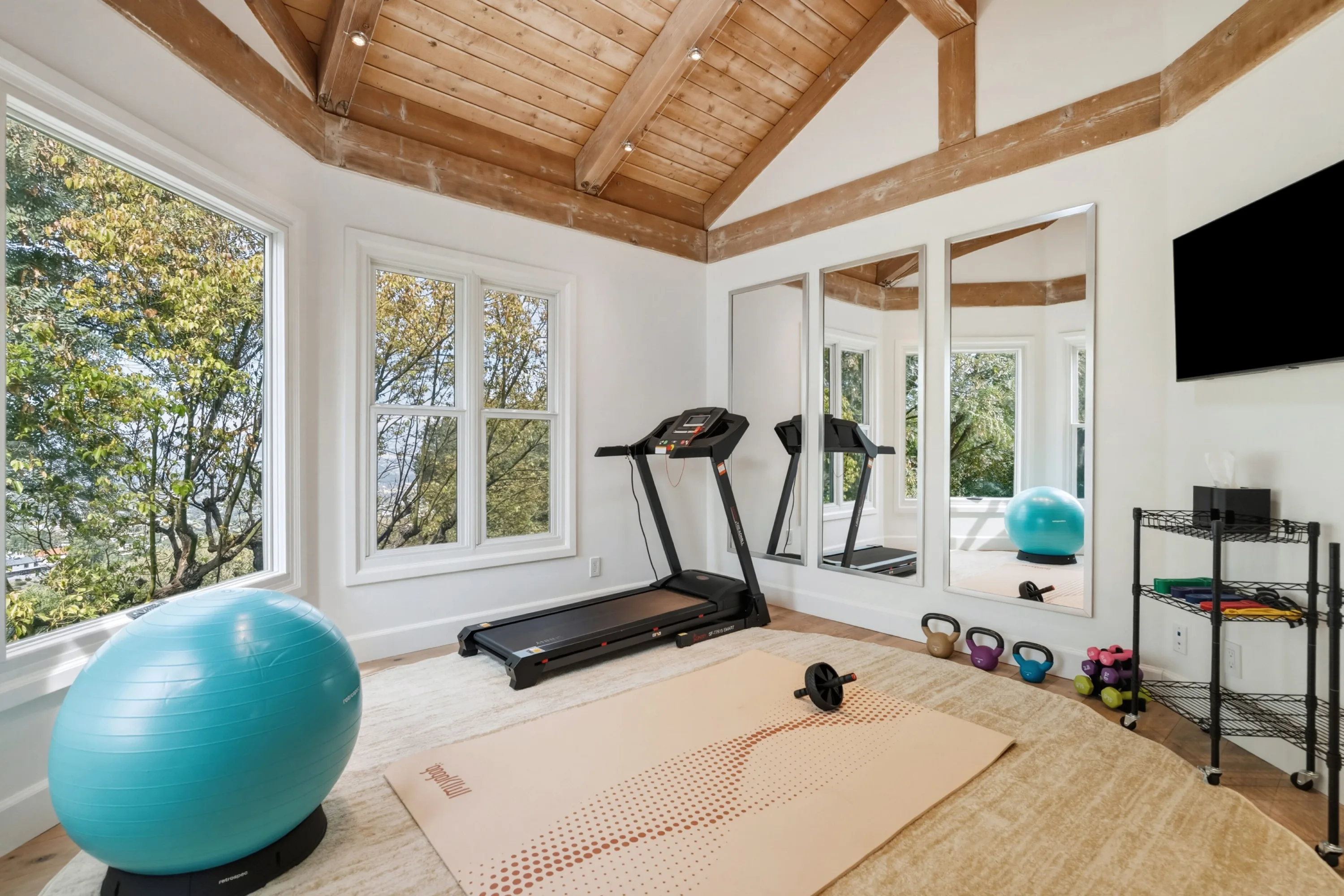 Home gym under a beamed vaulted ceiling with treadmill, exercise balls, kettlebells and mirrored wall