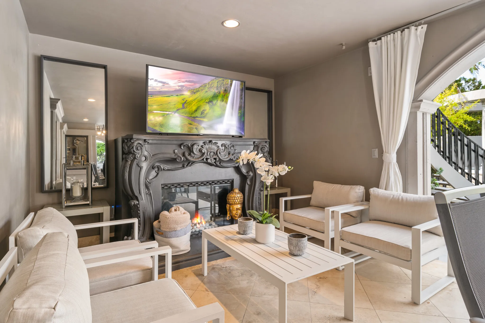 Sheltered terrace lounge with carved fireplace, wall-mounted TV and cream outdoor armchairs beside the pool arch
