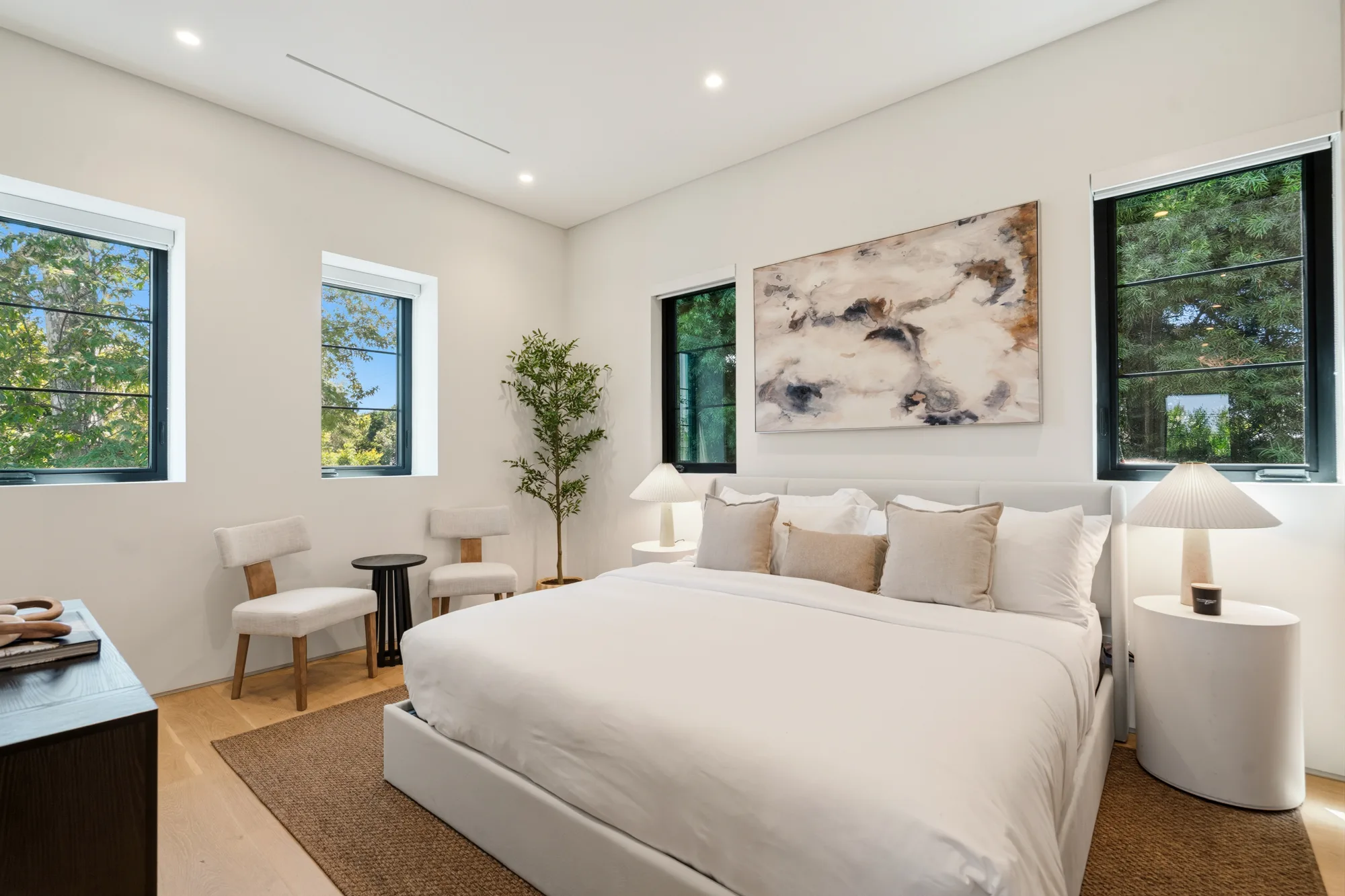 Primary suite in cream linens beneath an abstract canvas, with a seating nook and olive tree by the windows