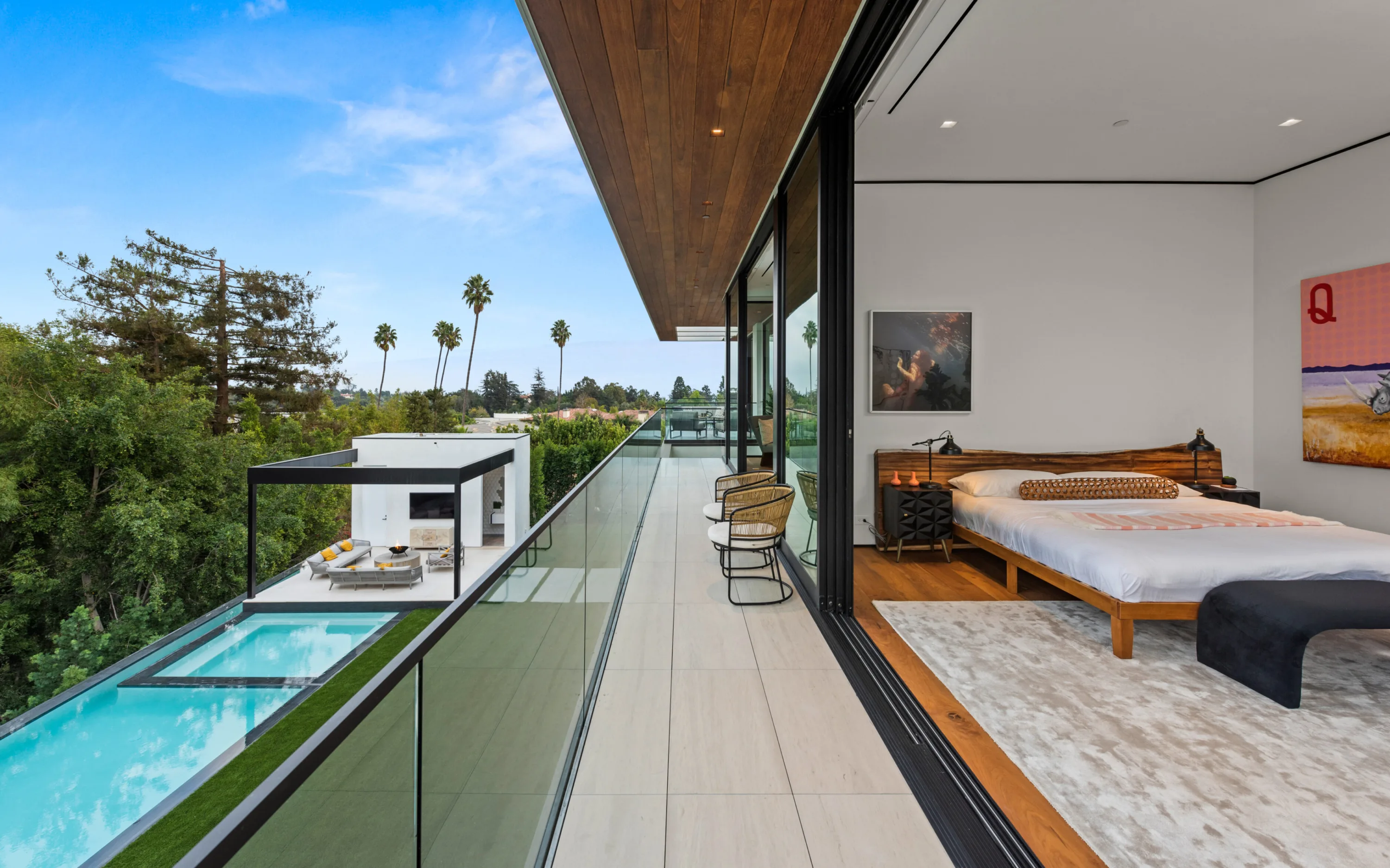 Bedroom opening to a glass-railed terrace overlooking the long pool and cabana, palms on the skyline