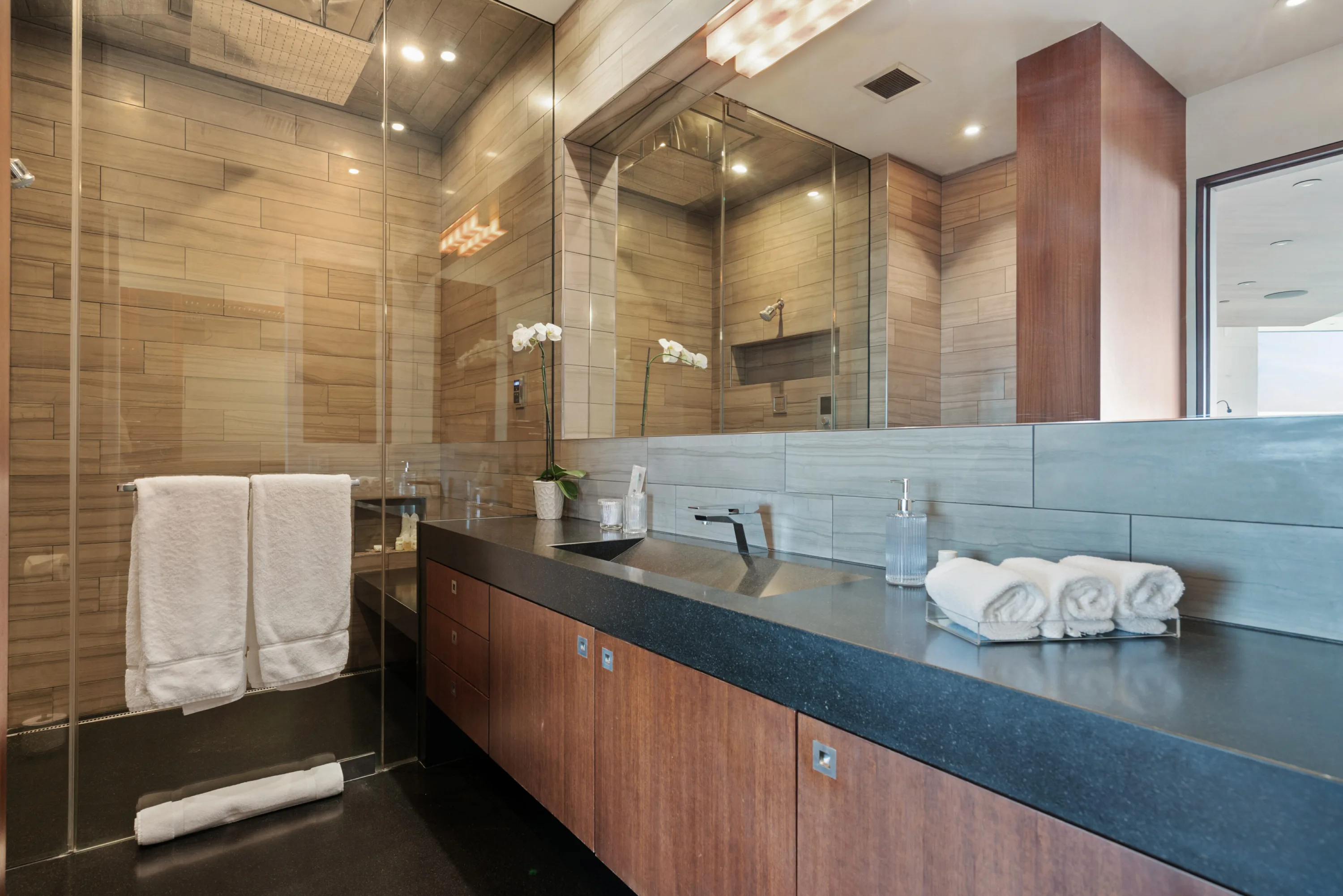 Stone-tiled bathroom with dark stone counter, orchid, rolled towels and a glass-enclosed shower
