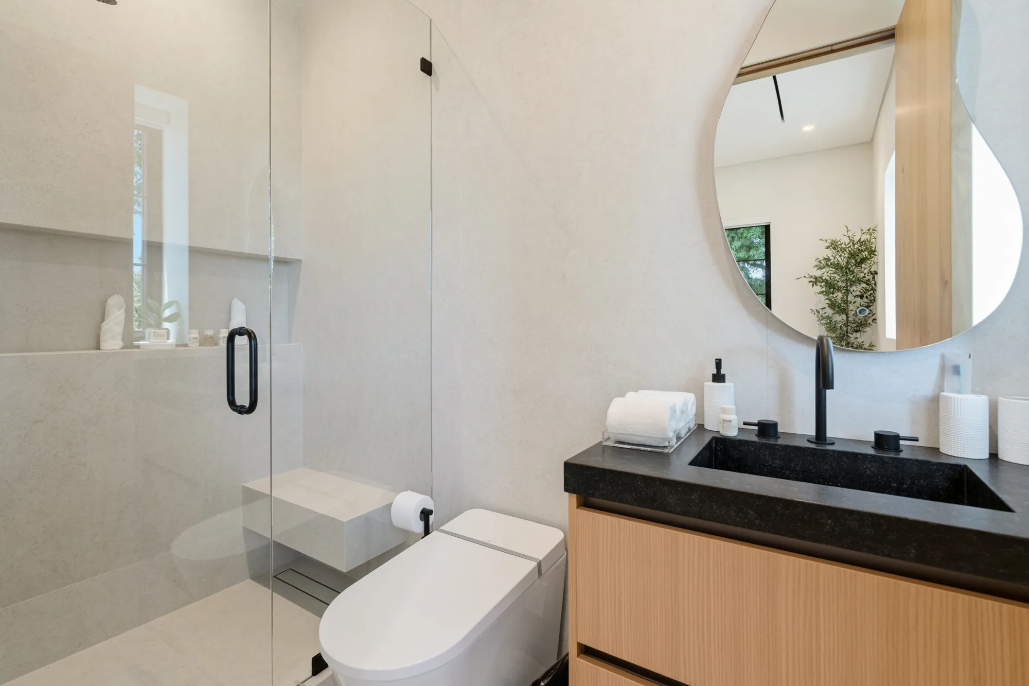 Compact ensuite with rounded wall mirror, black stone trough sink and glass-enclosed shower with bench
