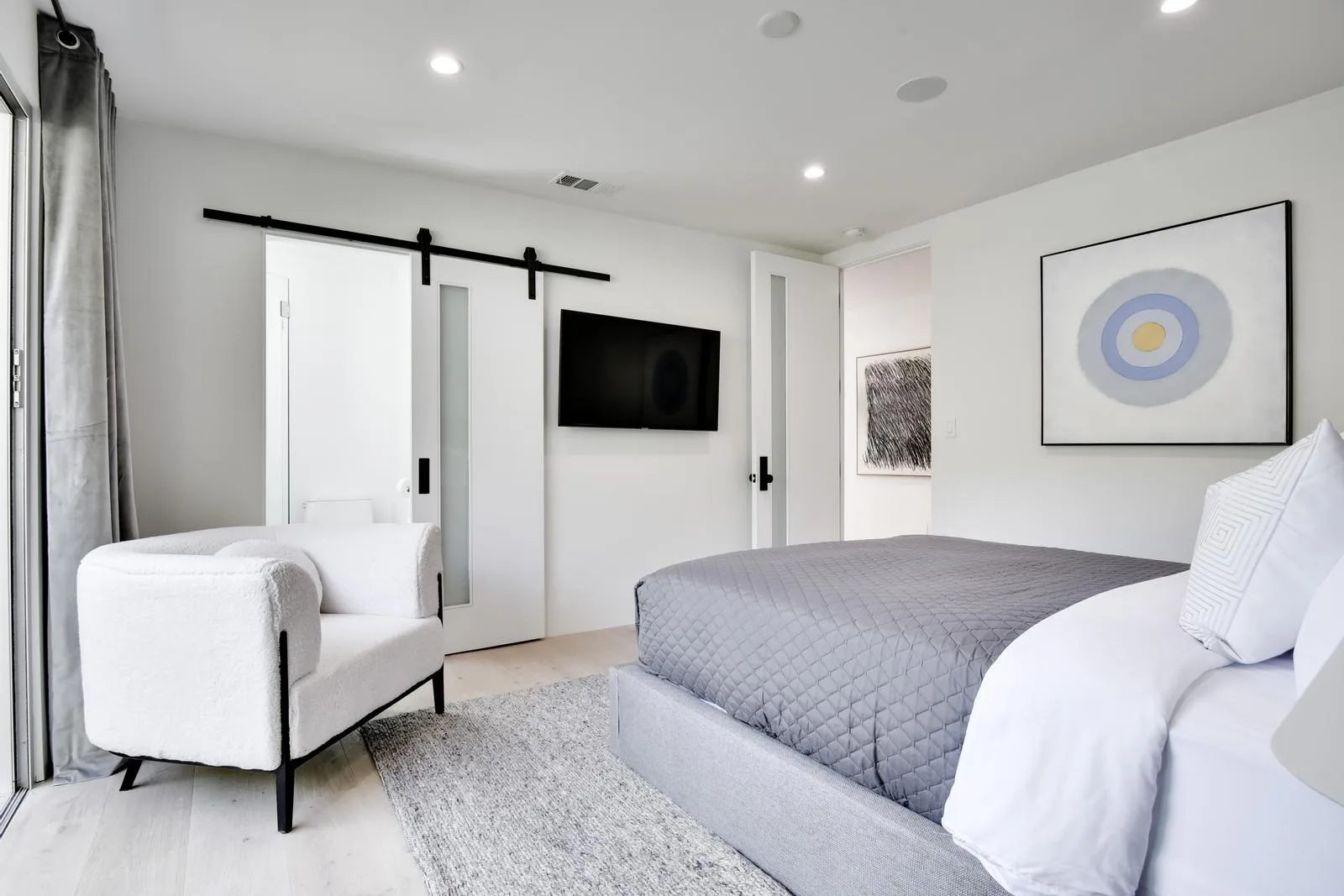 Crisp white guest room with grey quilted bed, bouclé armchair, barn-style sliding door and framed abstract art
