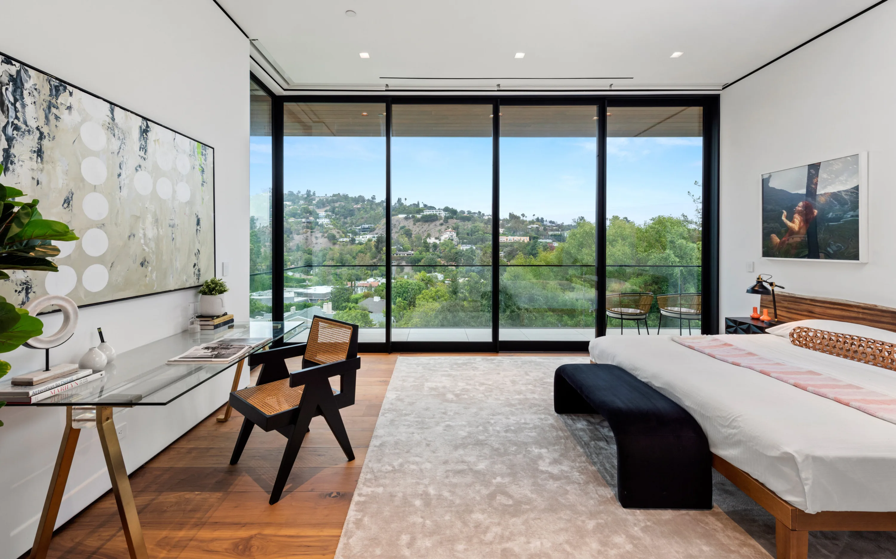 Guest bedroom with floor-to-ceiling glass framing green hillside homes, cane desk chair and velvet bench