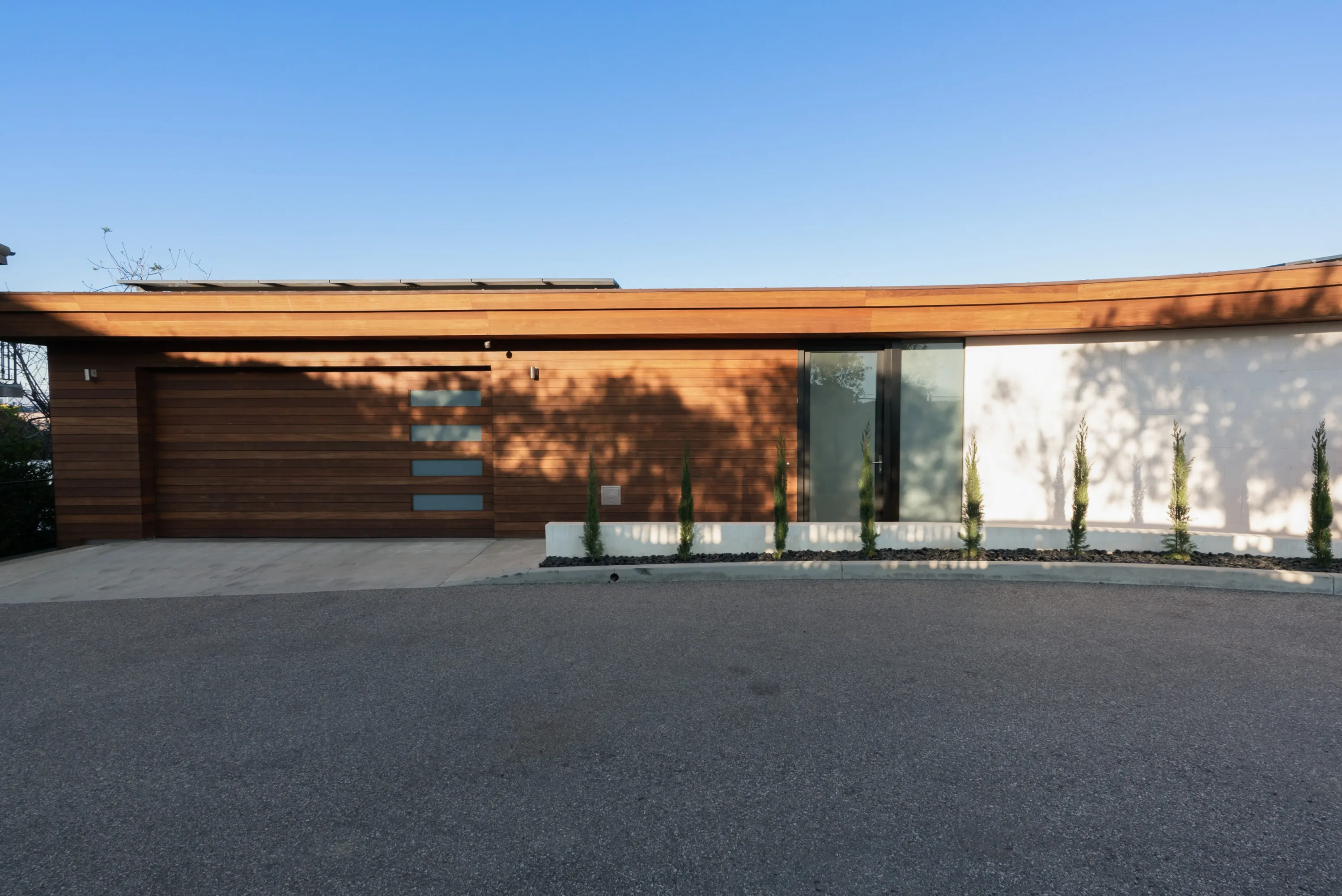 Street entrance with curved cedar roofline, walnut garage door and row of slim cypress