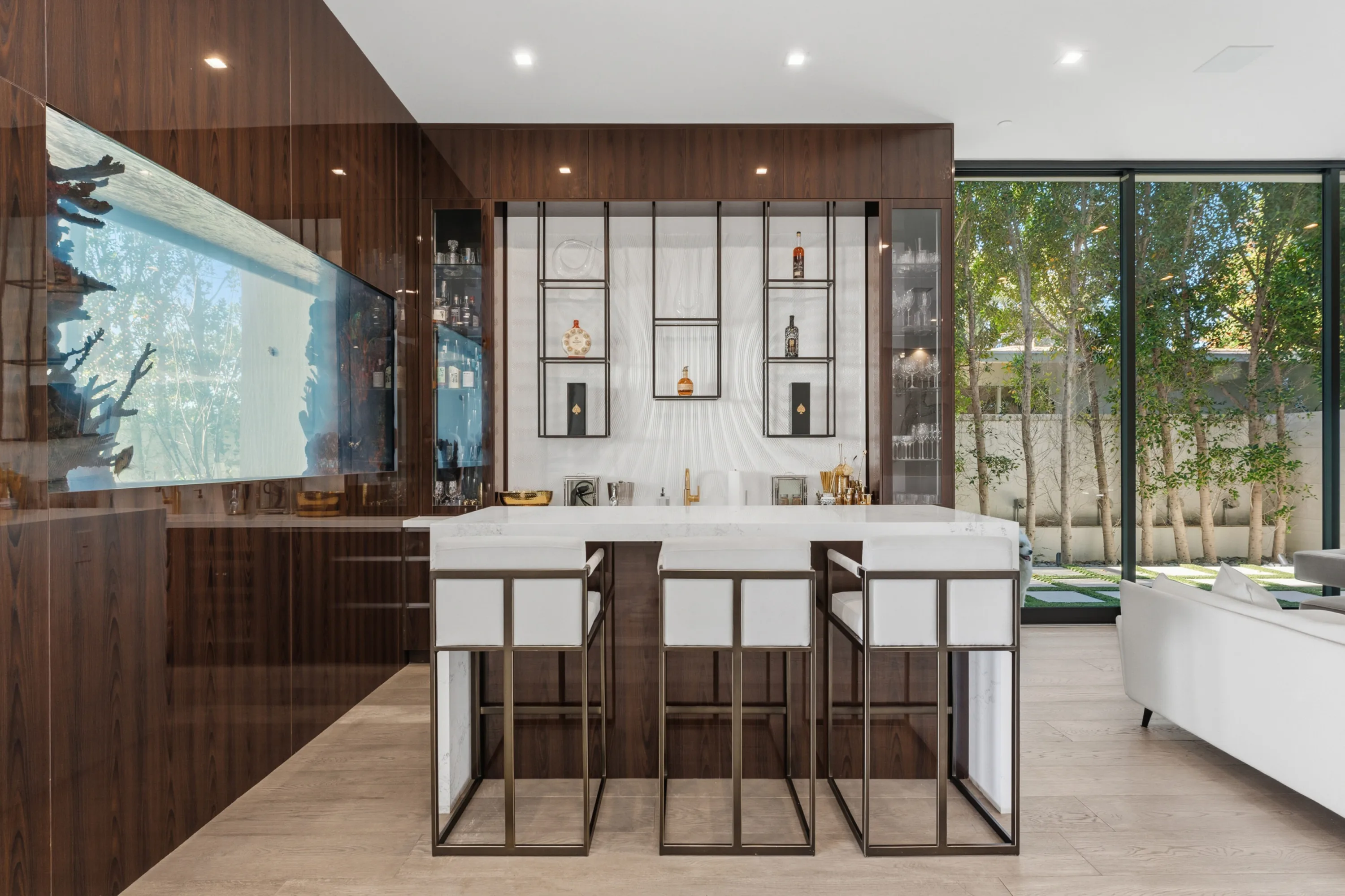 Walnut wet bar with white stone island, four barstools and a wall-set aquarium beside garden glazing