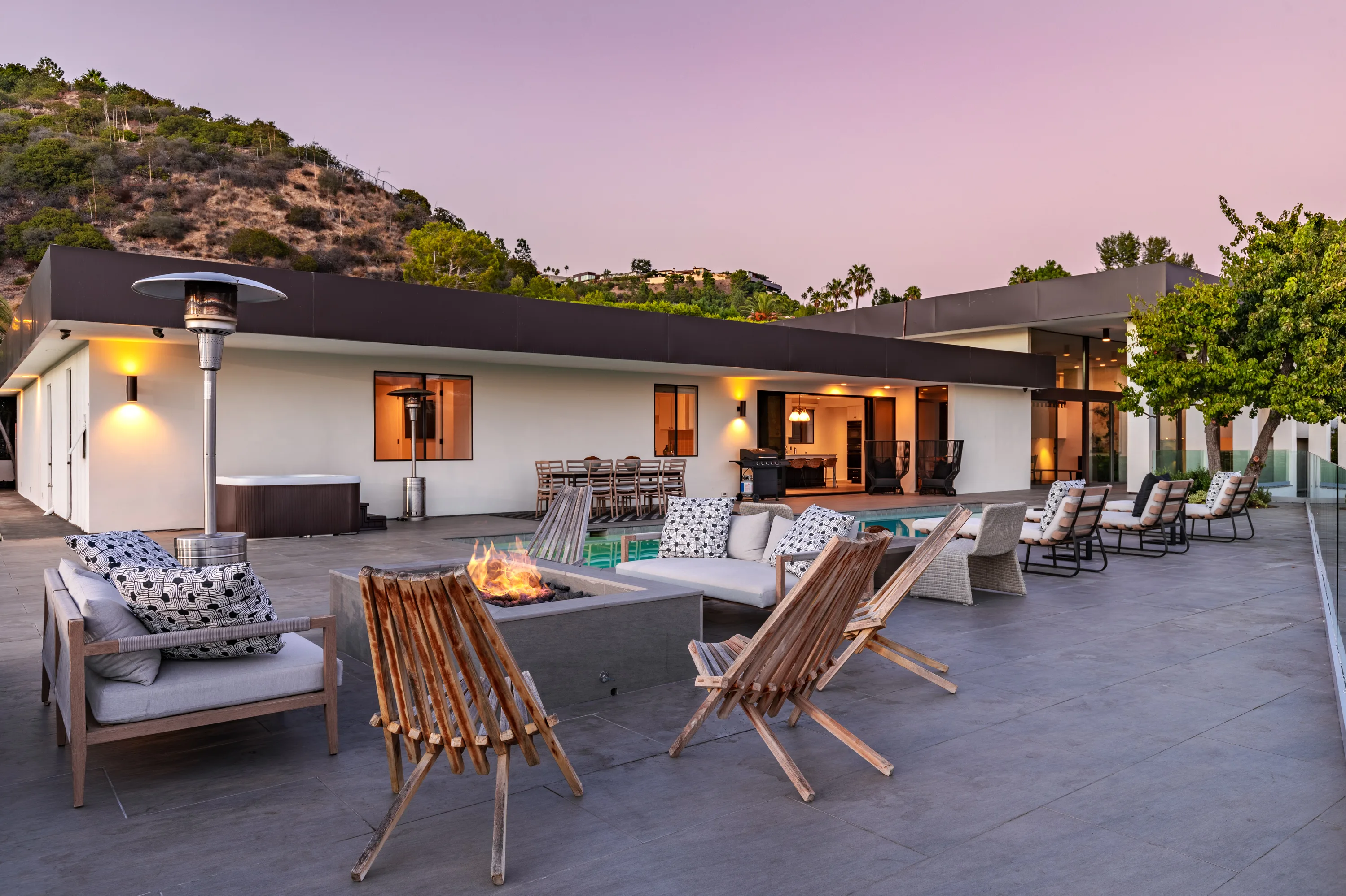 Poolside fire table and lounge chairs on the terrace, with spa, grill and glowing house at twilight