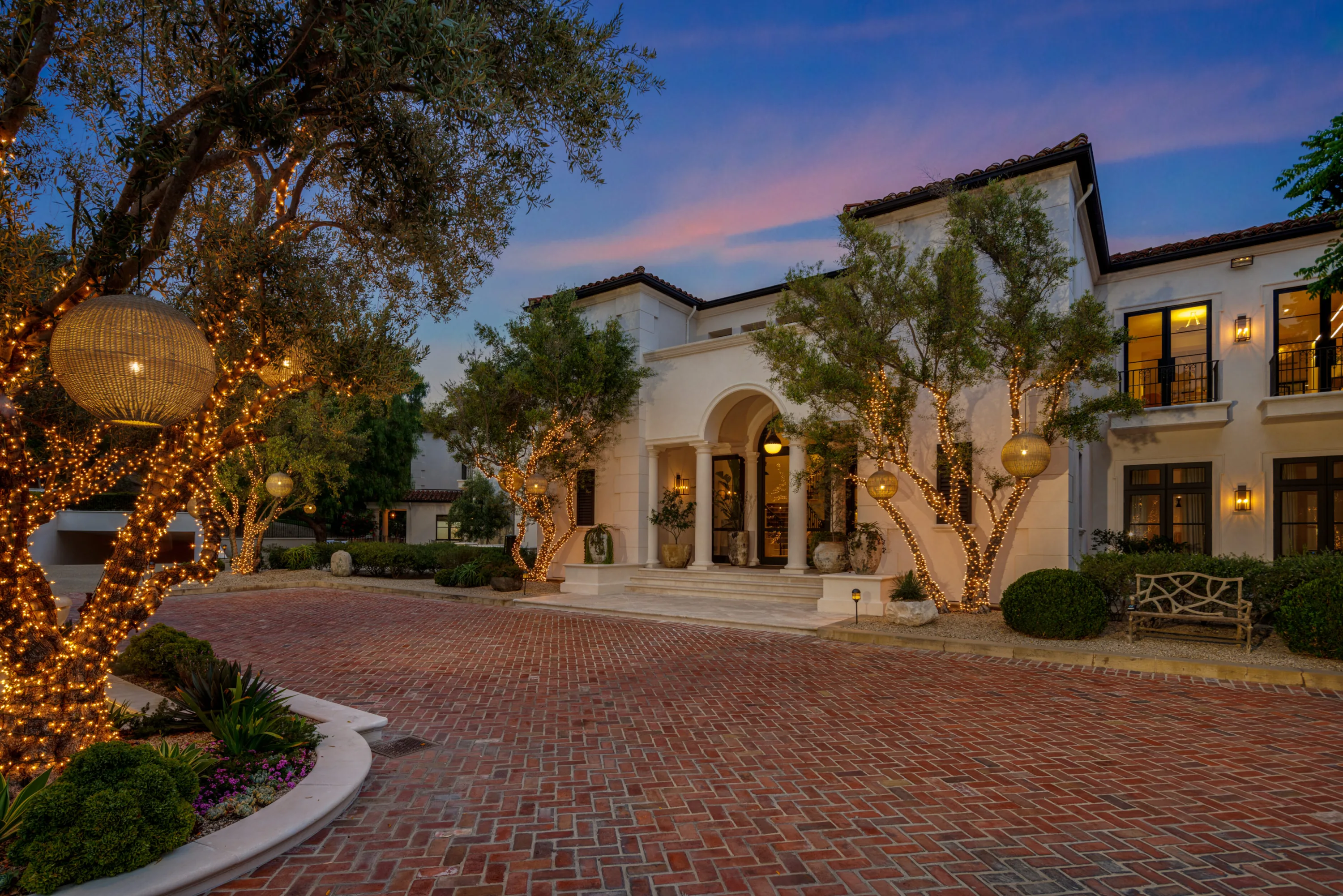 Arched front entrance and herringbone brick drive glowing under lantern-hung olive trees at sunset