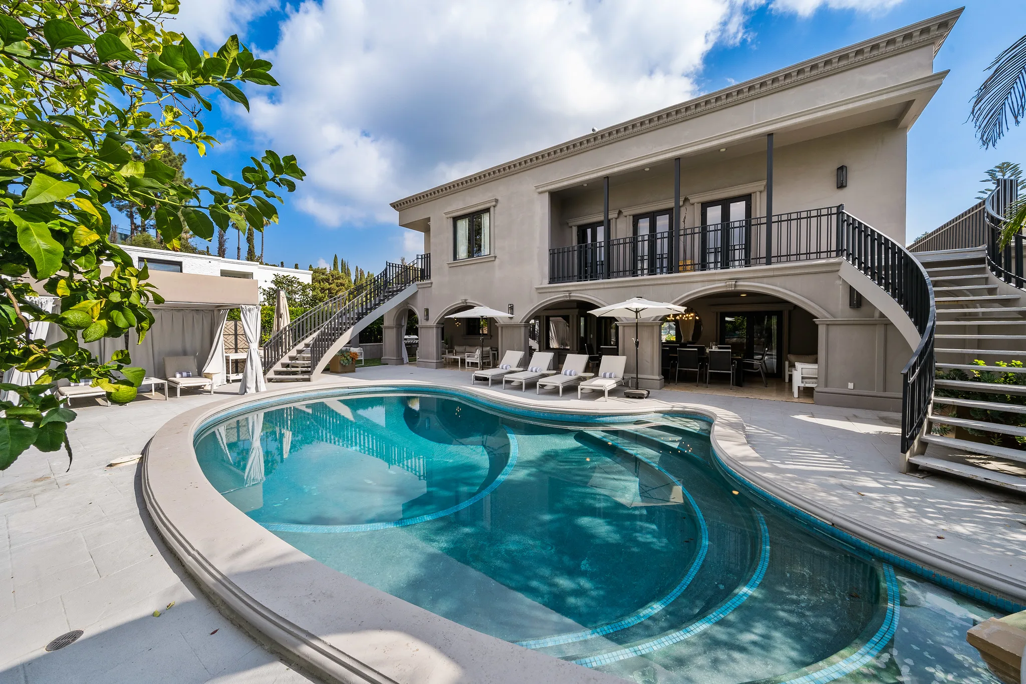 Curved pool and sun loungers below the rear facade, with twin staircases sweeping up to the first-floor balcony