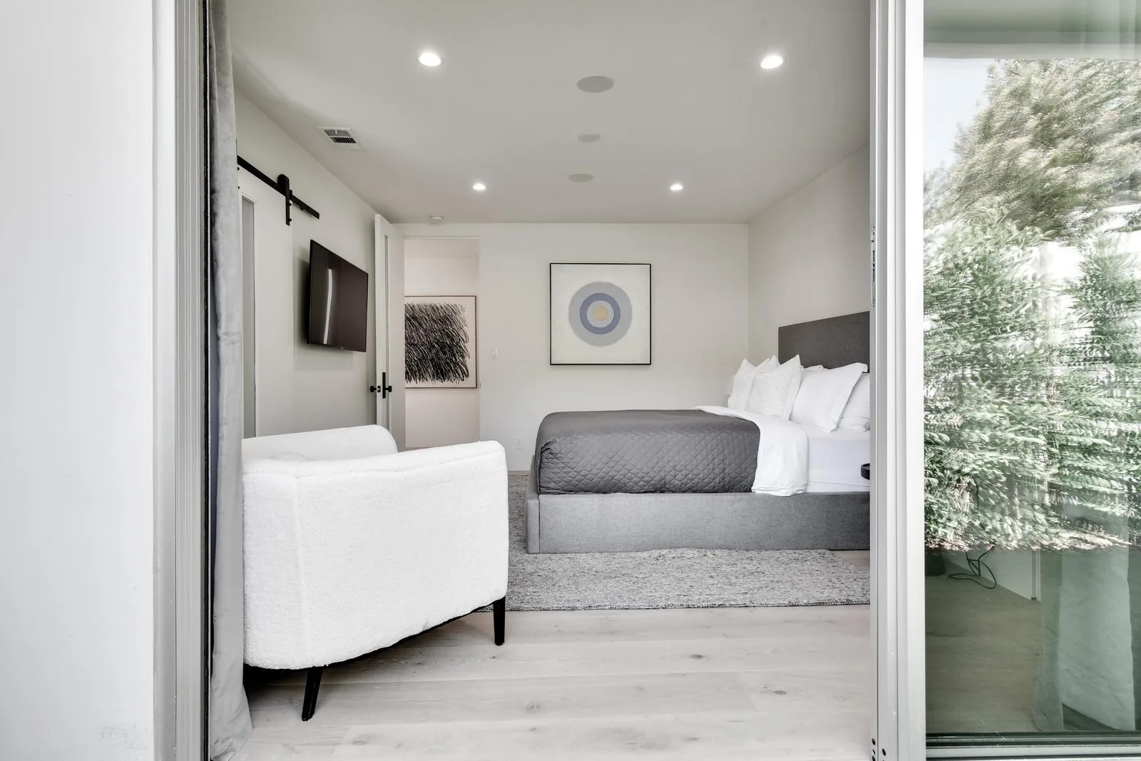 View from the terrace through open sliding doors into a grey-and-white bedroom with armchair and TV