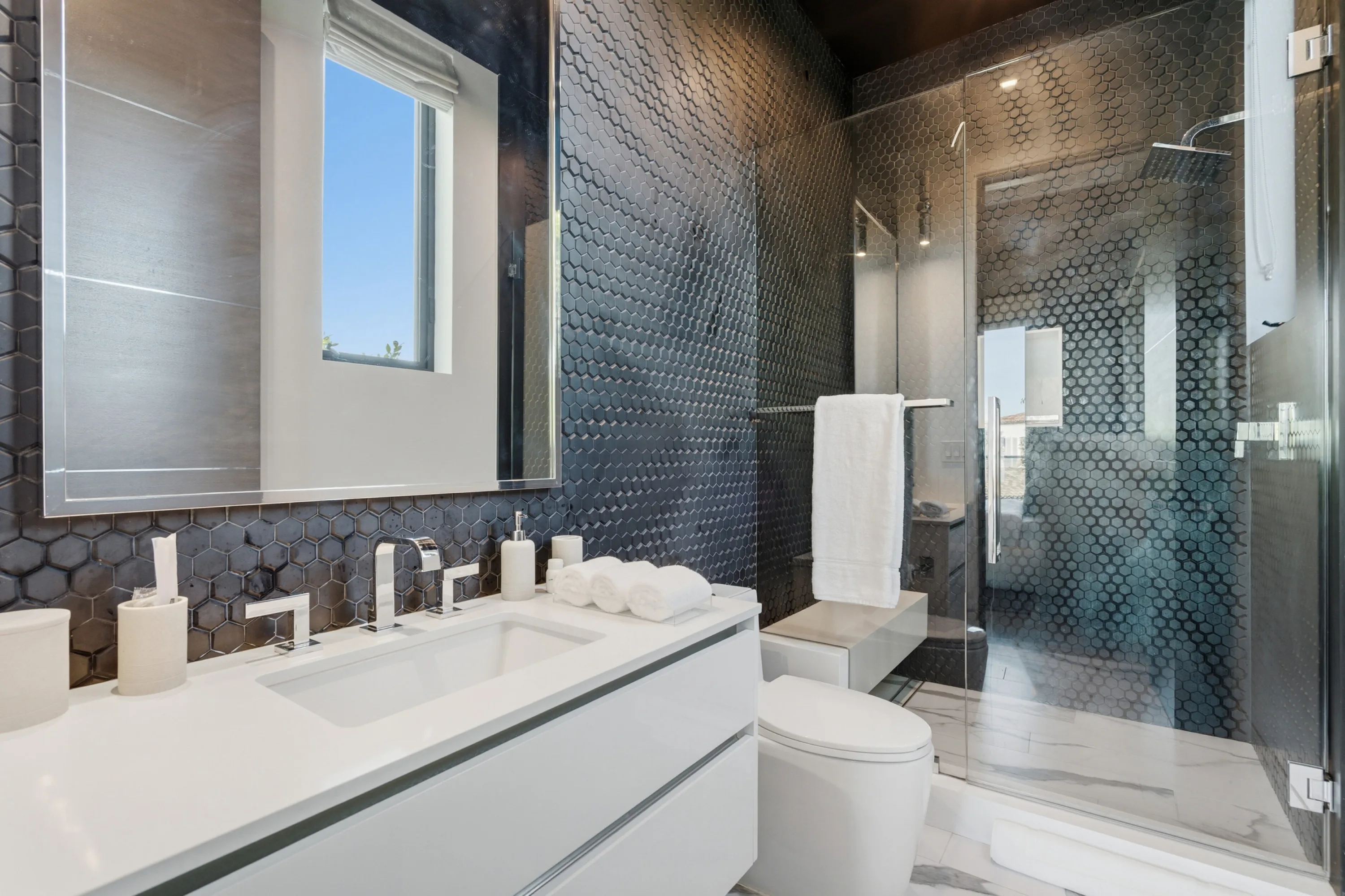 Guest bath clad in dark hexagonal mosaic with glass shower, white quartz vanity and rolled towels