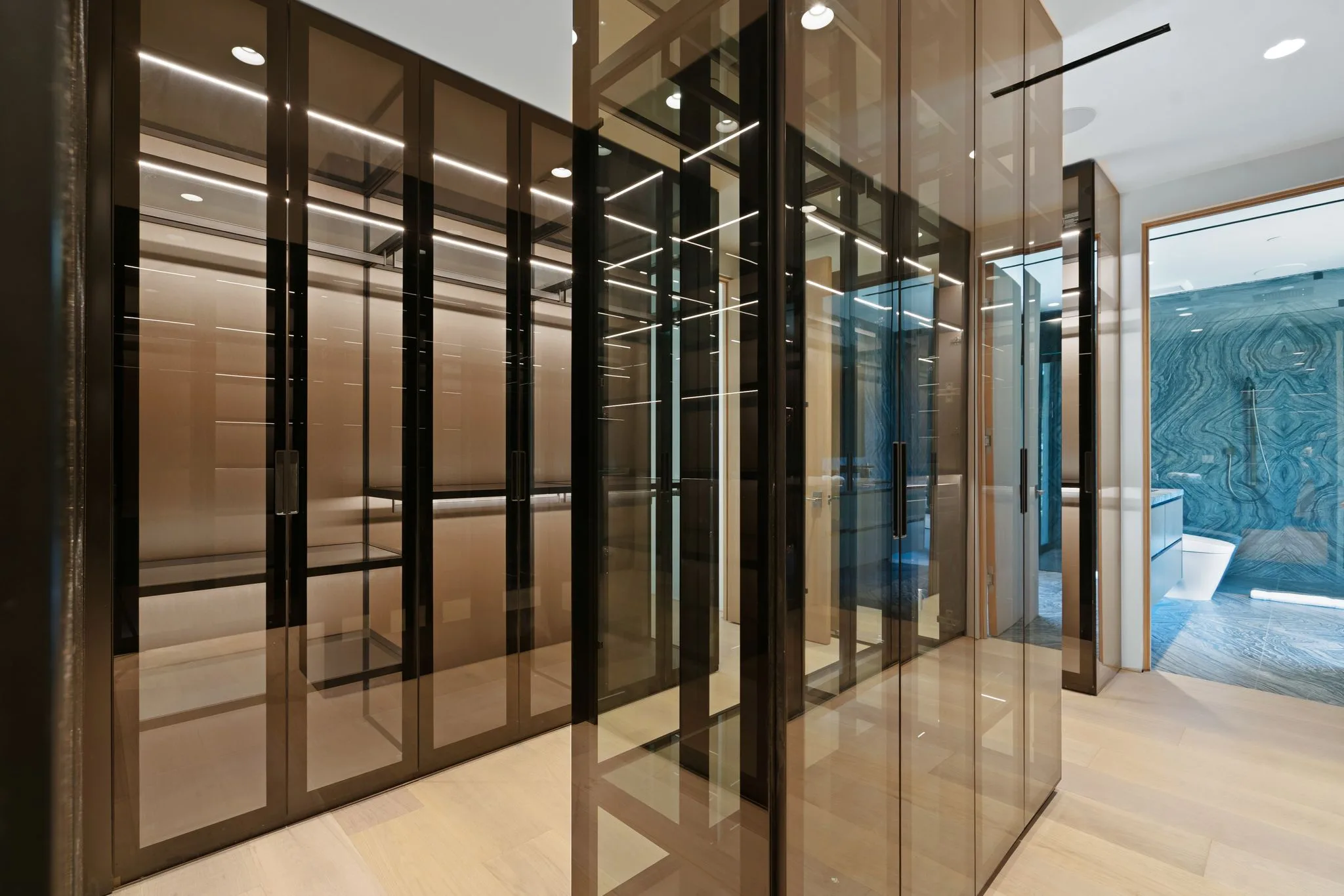 Bronze-tinted glass wardrobe bays lining a lit dressing corridor, marble bathroom visible beyond