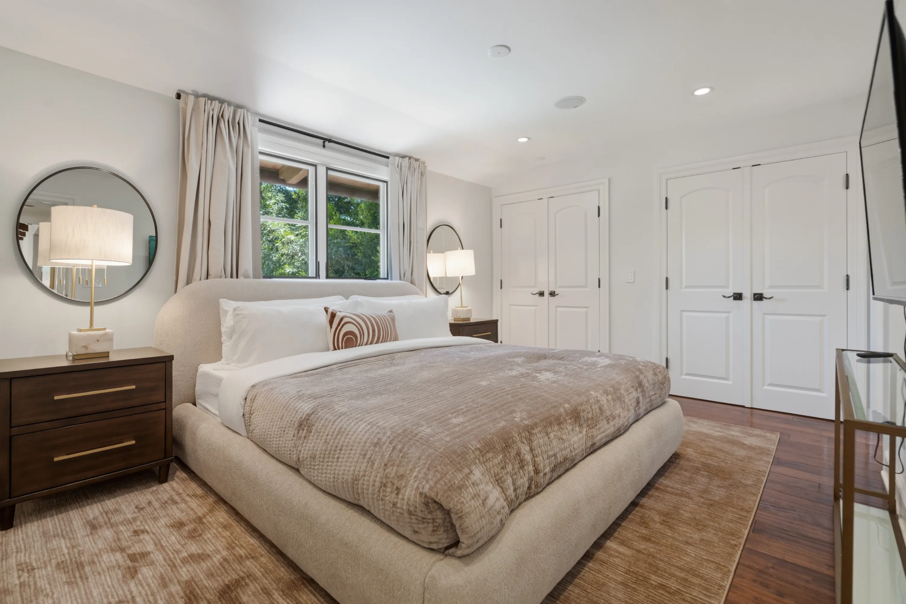 Bedroom in warm neutrals with round mirror, upholstered bed and garden window above the headboard