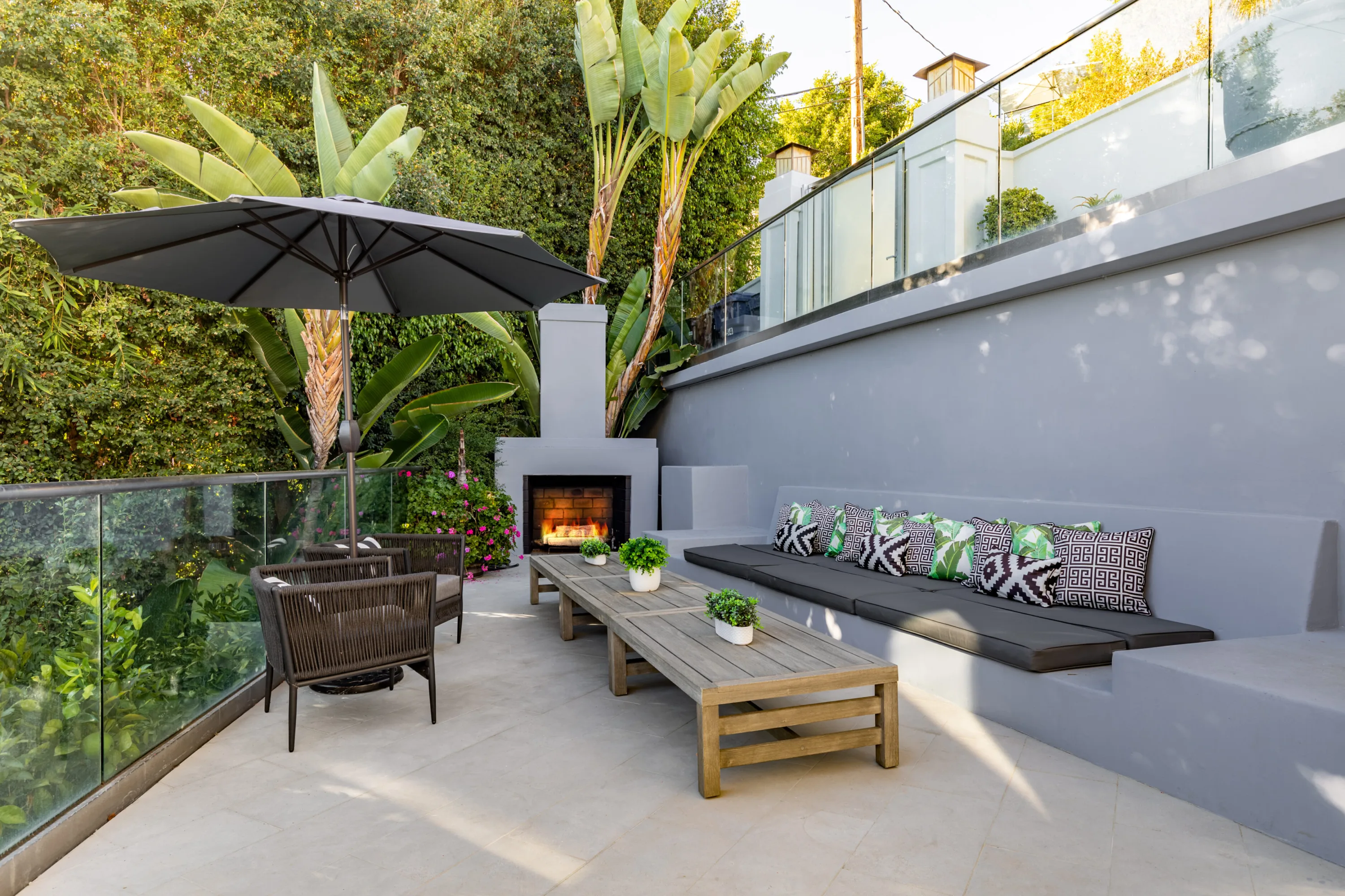 Sunken terrace with lit outdoor fireplace, cushioned banquette in palm-print pillows and shaded low tables