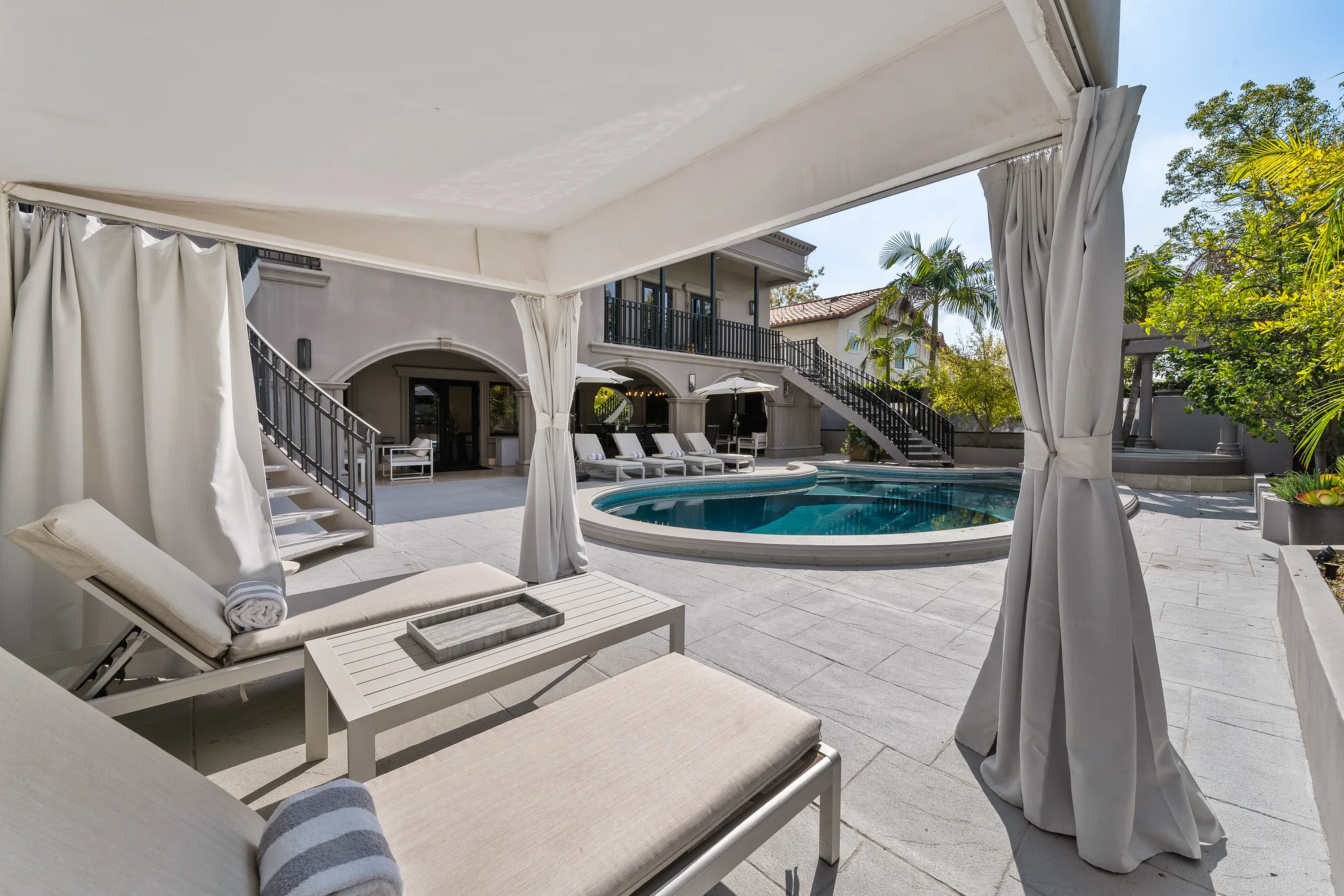 View from inside the curtained poolside cabana across loungers to the kidney-shaped pool and arcaded terrace