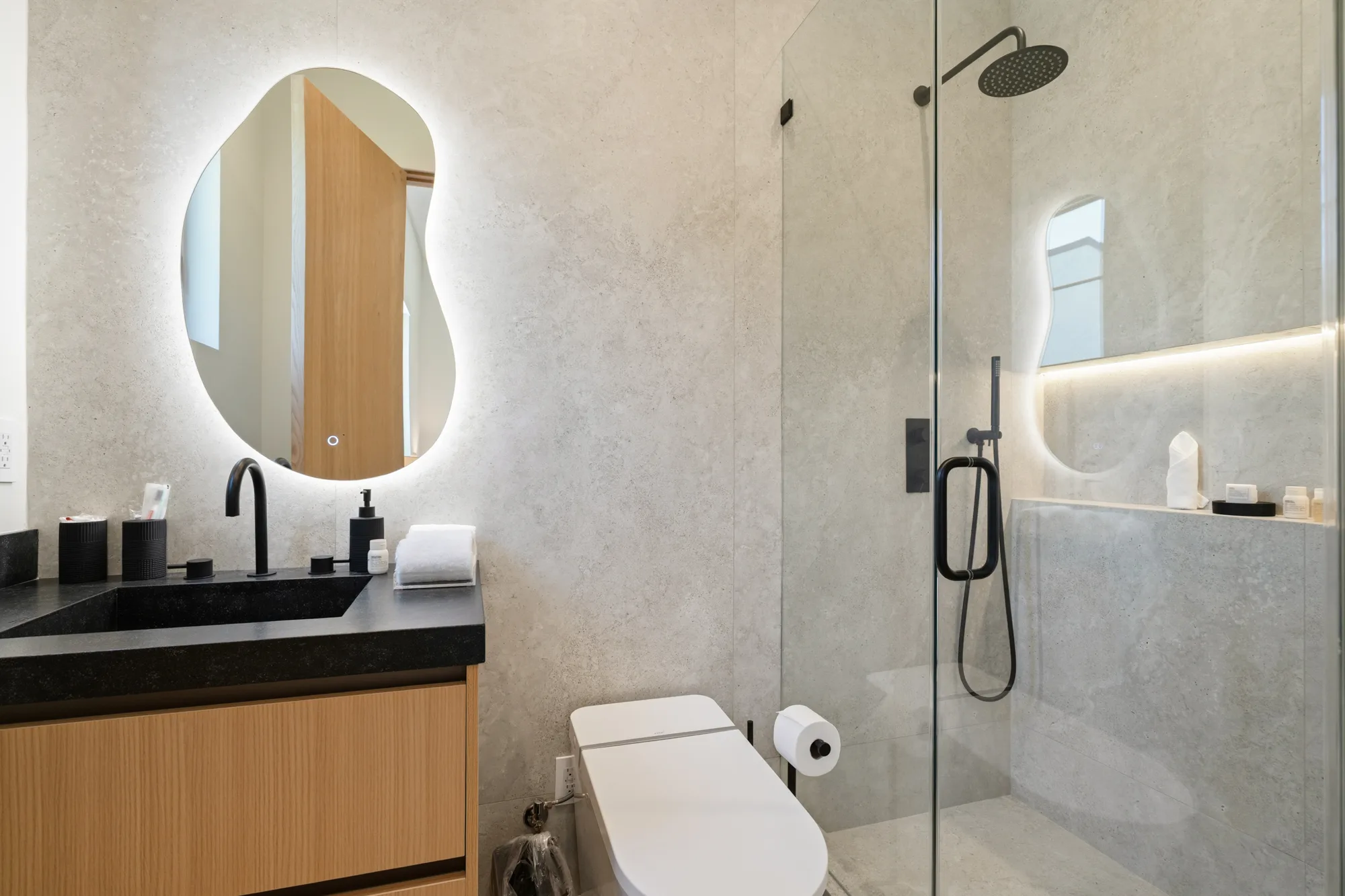 Third bathroom pairing a glowing freeform mirror with a matte black fittings and a lit shower niche