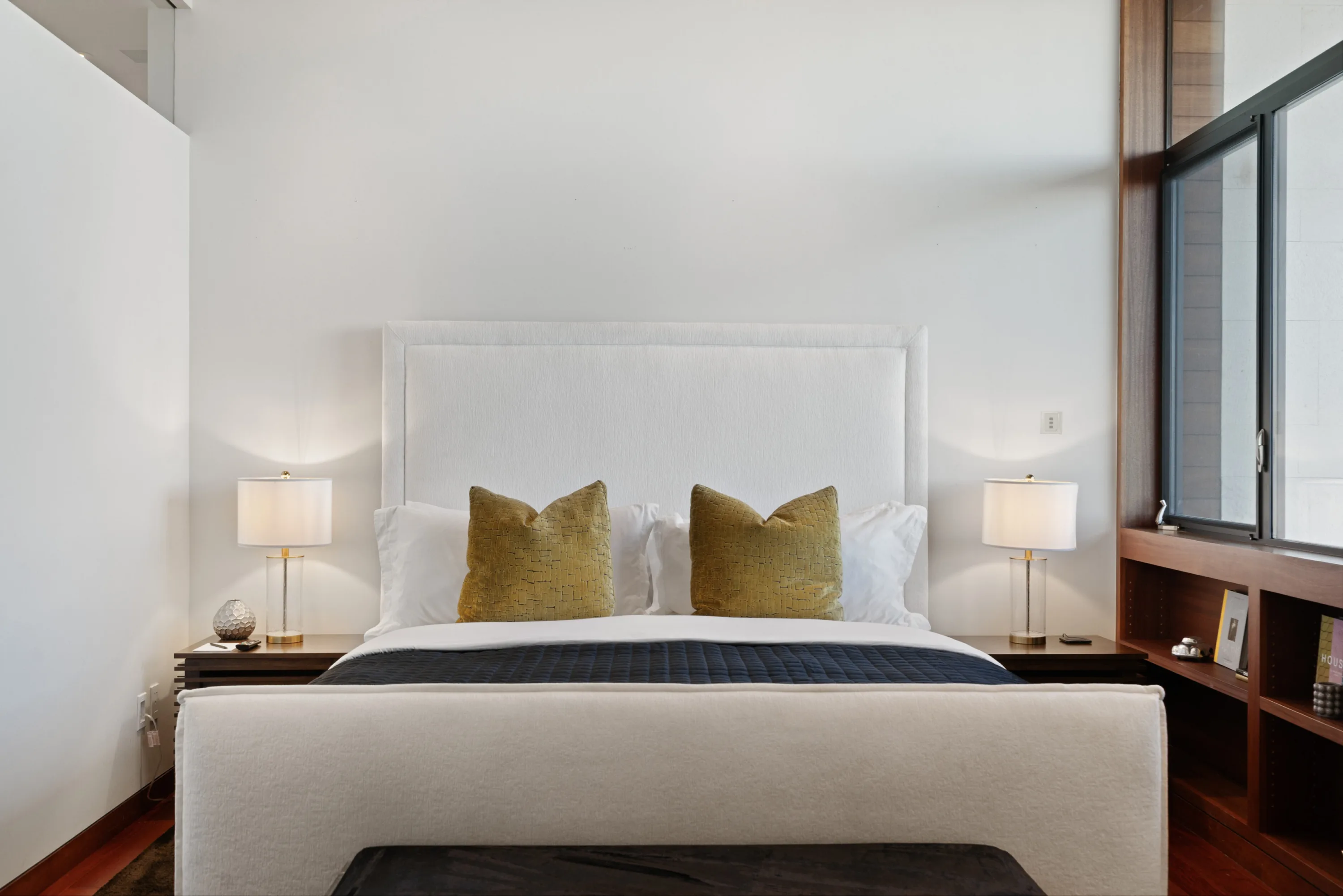 Head-on view of the same bedroom's tall white headboard, gold velvet cushions and matching bedside lamps