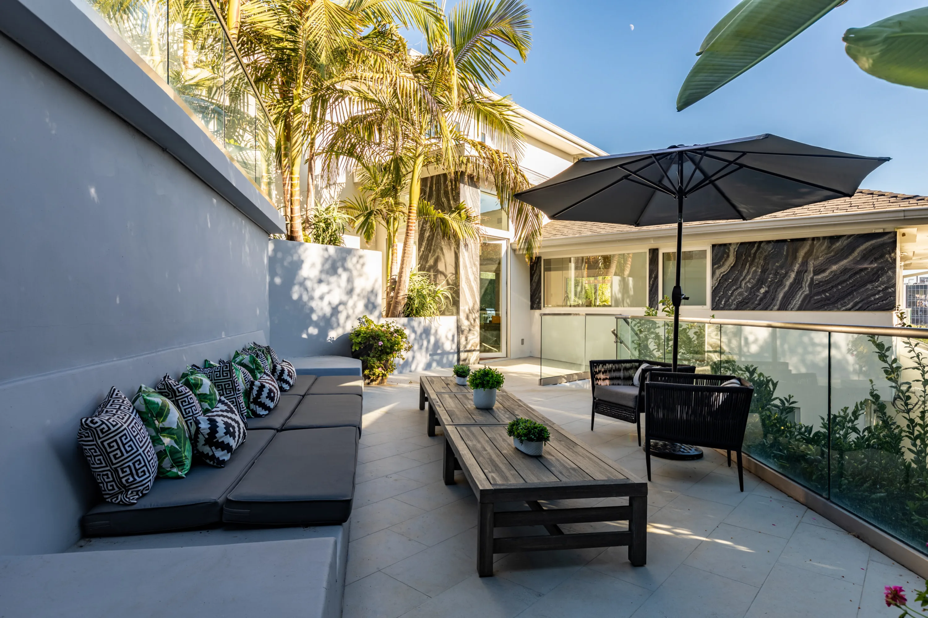 Glass-railed courtyard lounge with built-in bench seating and parasol, palms rising against the villa's facade