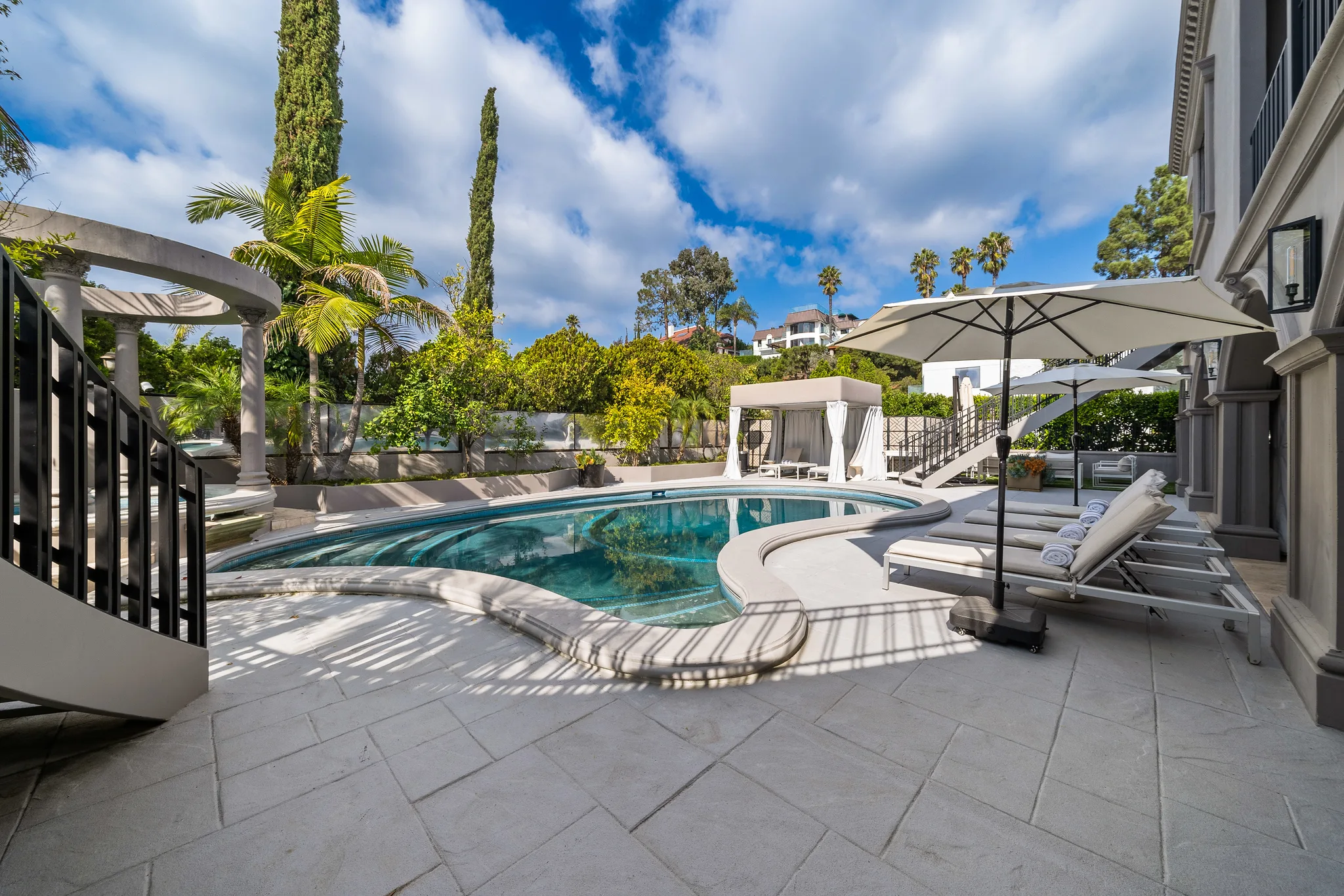 Sunlit pool deck with shaded loungers, palms and cypress framing the Hollywood Hills ridgeline beyond