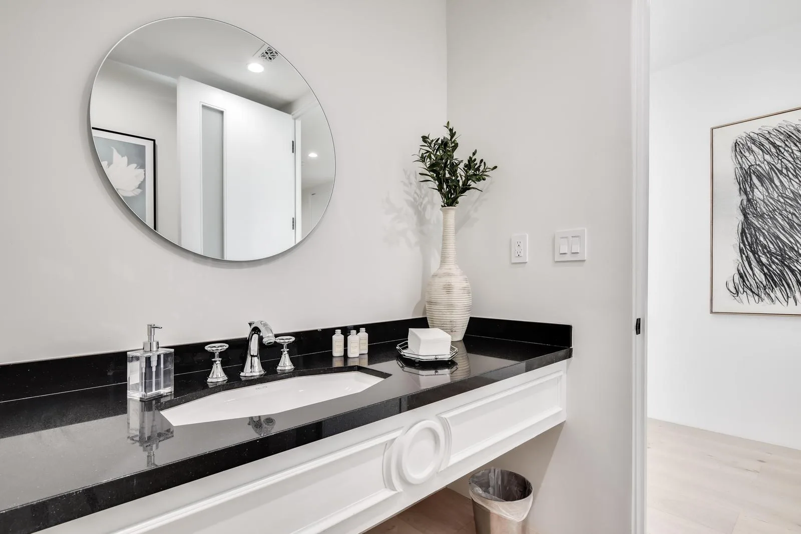 Powder room with black stone counter, round mirror and toiletries, opening onto a hall hung with abstract art