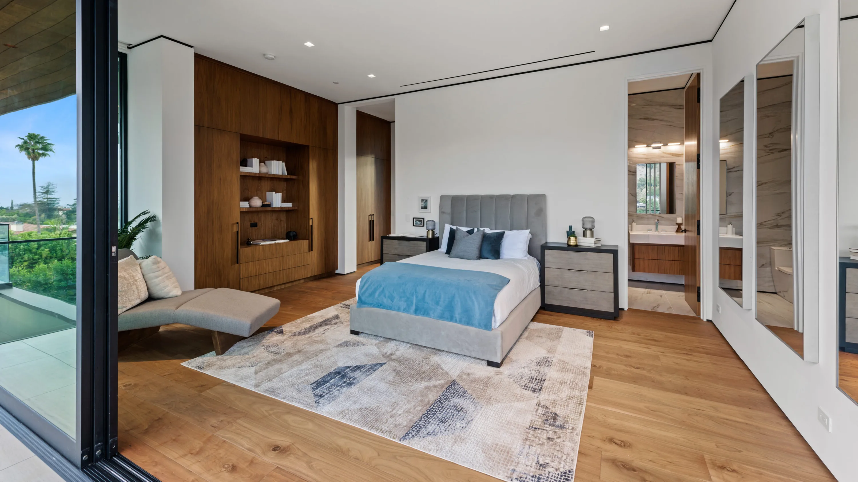 Bedroom in soft blue with built-in walnut joinery, chaise by the sliding glass and en-suite beyond