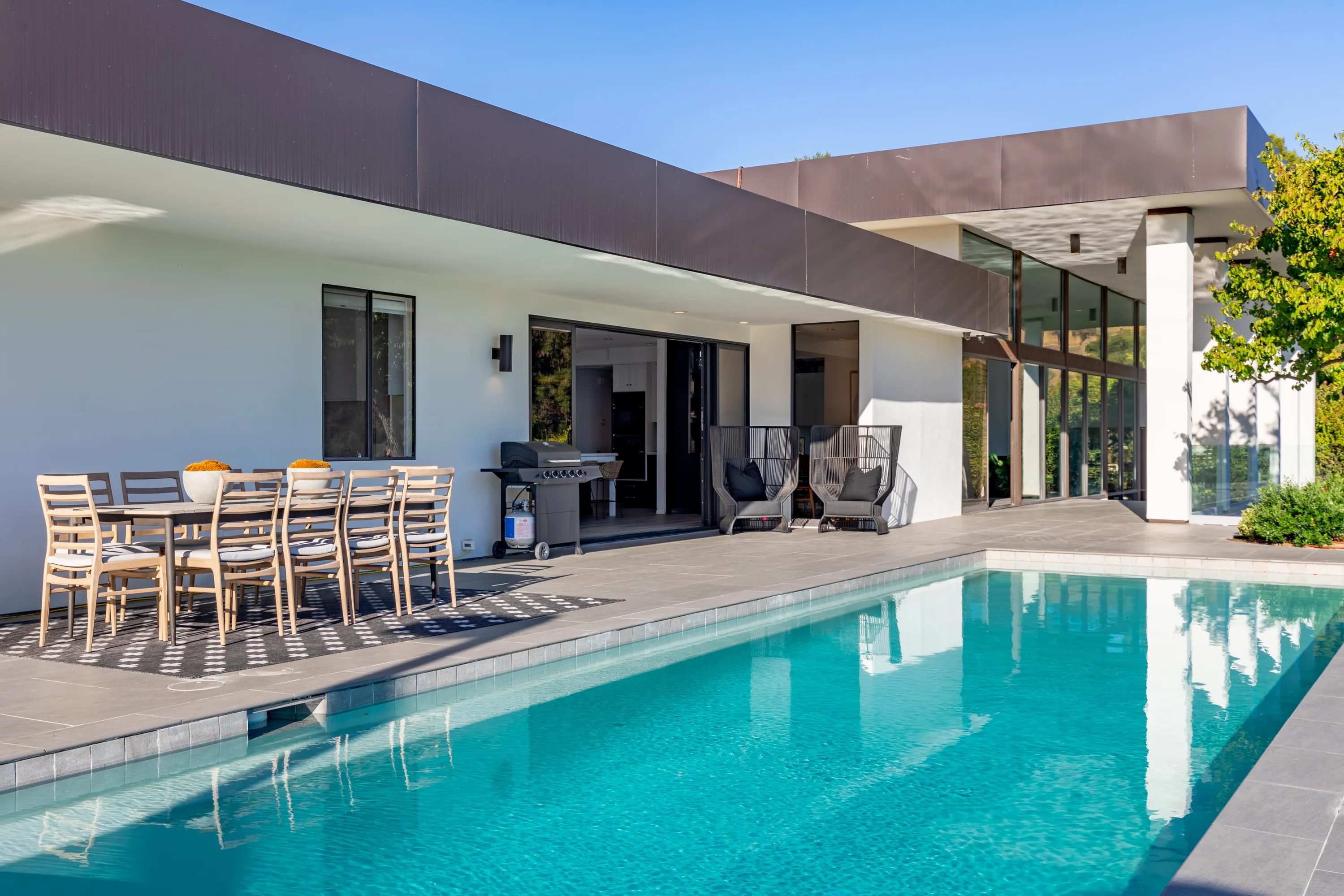 Poolside terrace with long dining table, grill and armchairs under the deep overhang