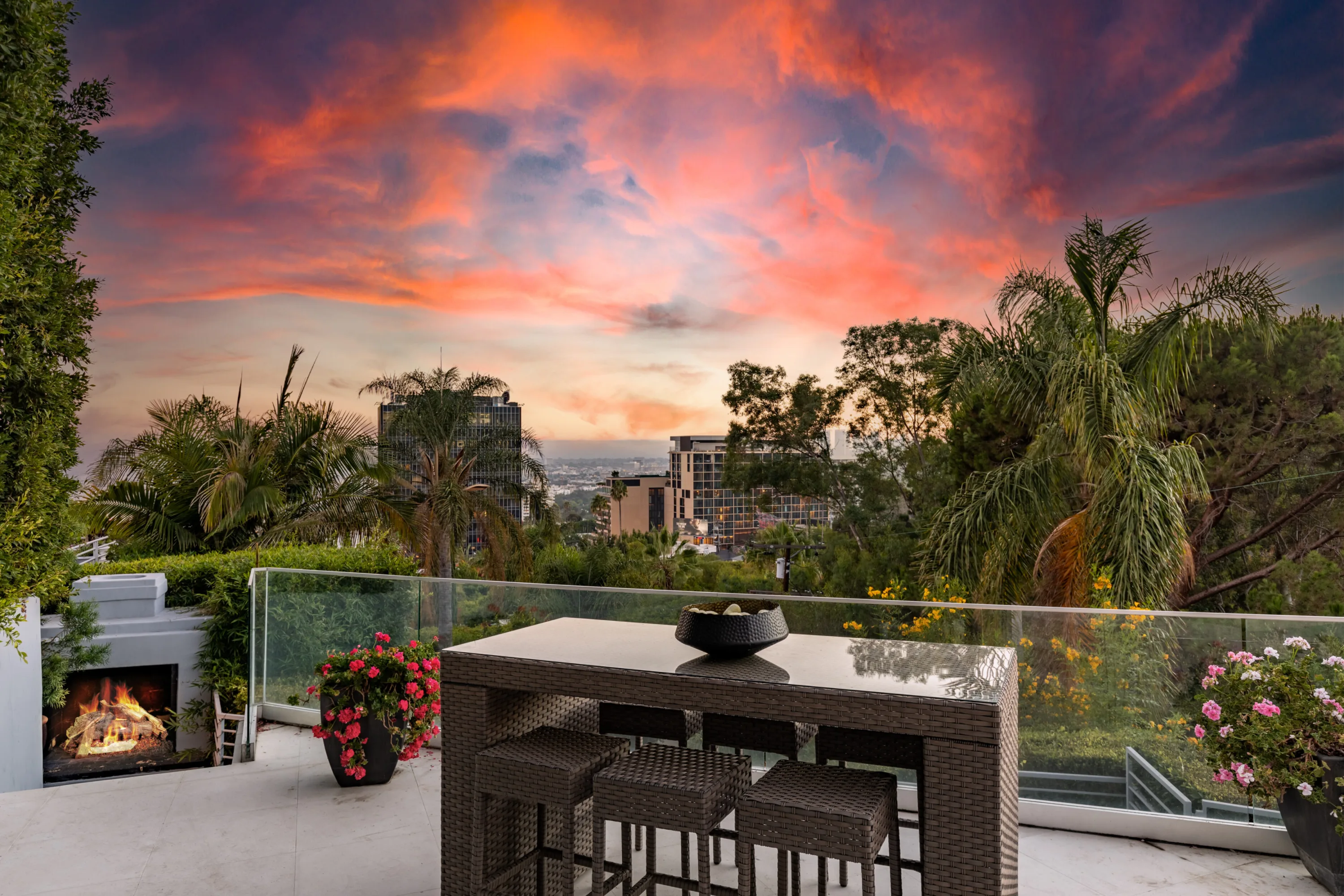 High-top bar table and stools on the terrace, city skyline and palms under a blazing pink sunset